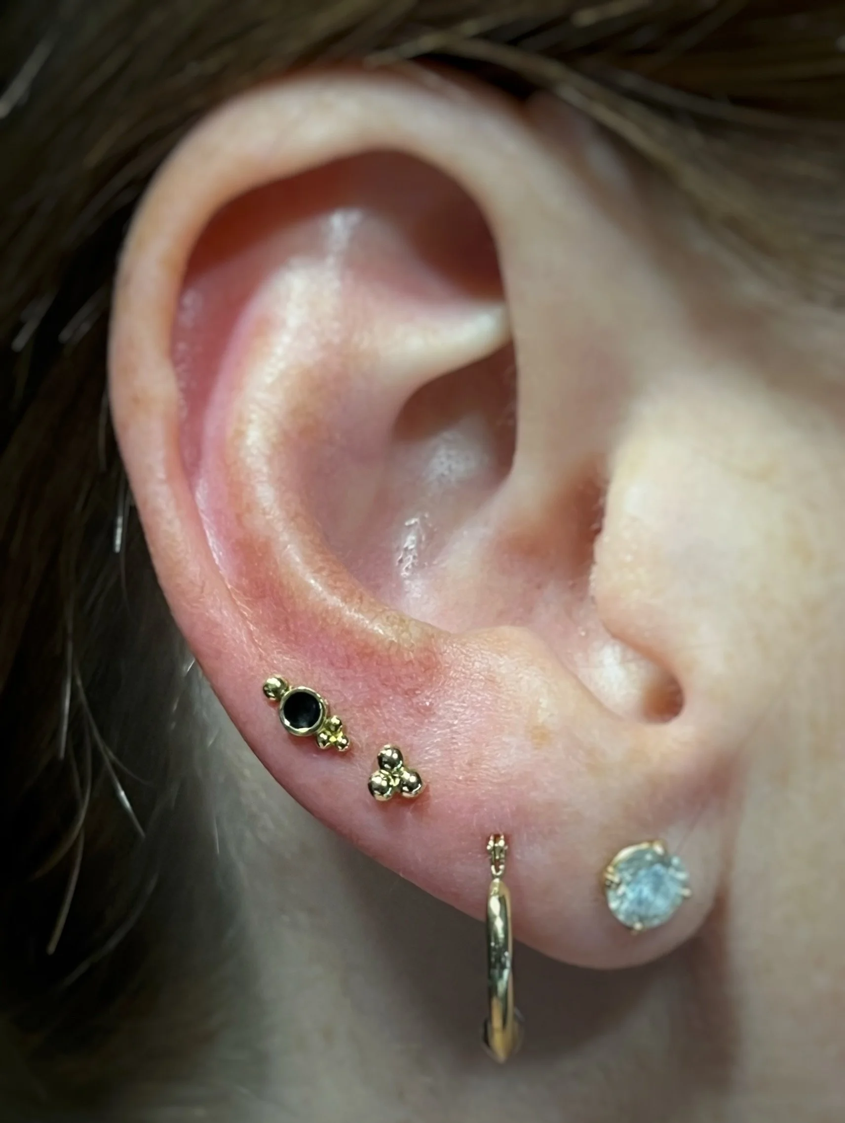 Close-up of a person's ear with multiple gold piercings, including a varicolored gemstone dermal piercing at the earlobe and a gold hoop with a dangling charm.