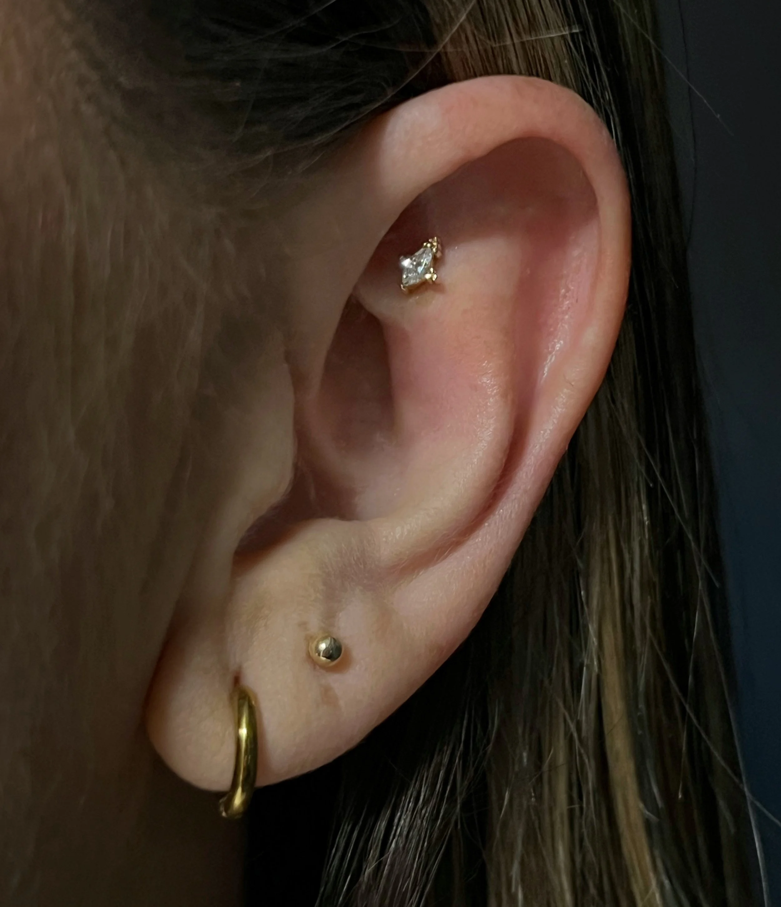Close-up of a person's ear with a small gold stud earring featuring a clear gemstone and a gold hoop earring, with long, dark hair partially visible.