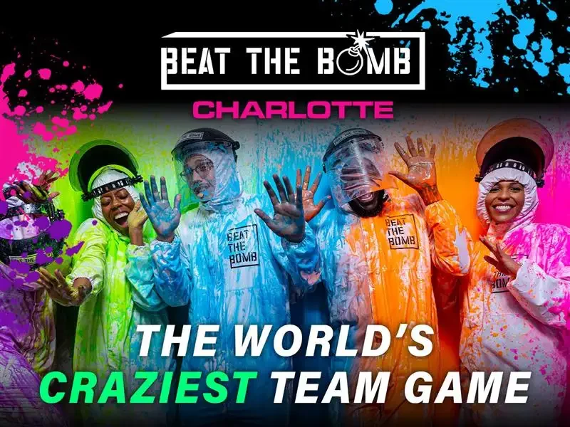 Group of five women in colorful protective suits and helmets, smiling and waving, with splashes of vibrant paint around them, promoting a team game called 'Beat the Bomb' led by Charlotte.