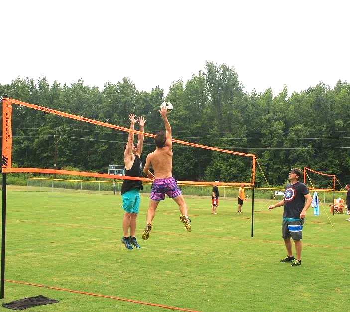 Two men are playing volleyball on a grassy field, with one jumping to hit the ball and the other trying to block, while other people are visible in the background in an adult volleyball league in Charlotte NC