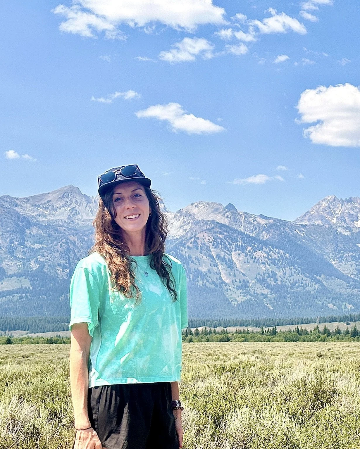 A woman standing in a grassy field with mountains and a blue sky with scattered clouds in the background.
