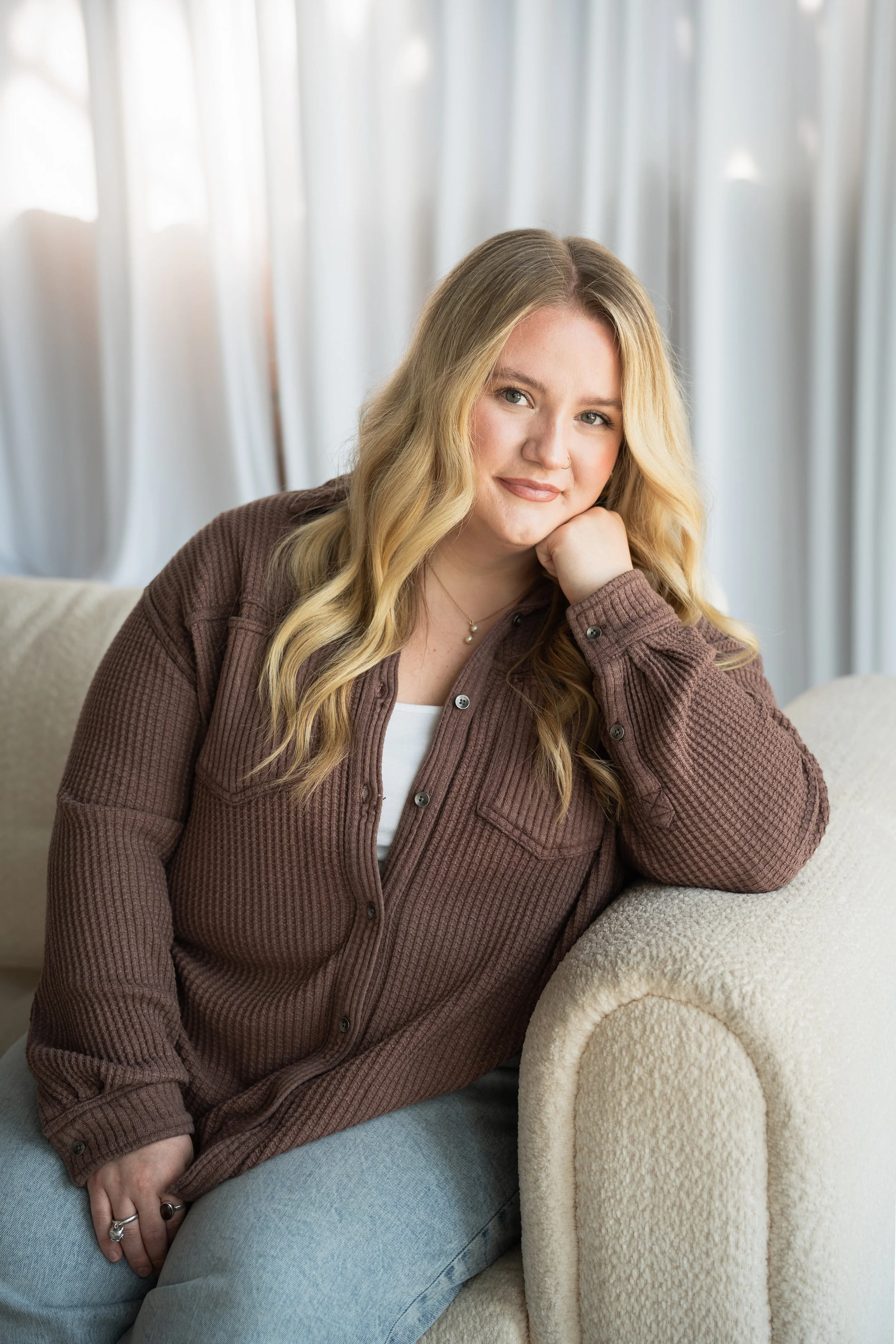 A young woman with blonde wavy hair, sitting and smiling at the camera, wearing a black long-sleeve shirt and black pants against a plain white background.