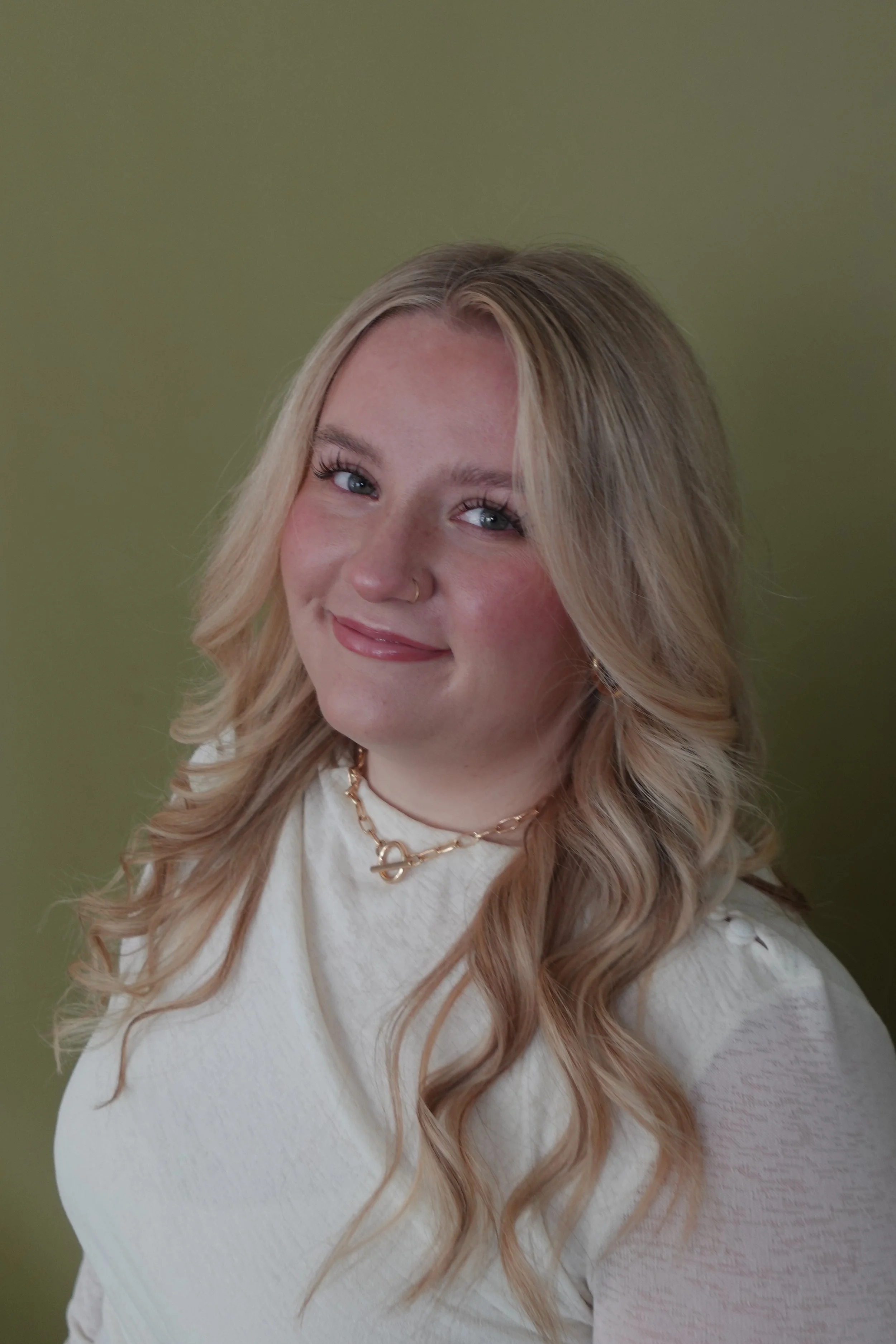 A young woman with blonde wavy hair, sitting and smiling at the camera, wearing a black long-sleeve shirt and black pants against a plain white background.