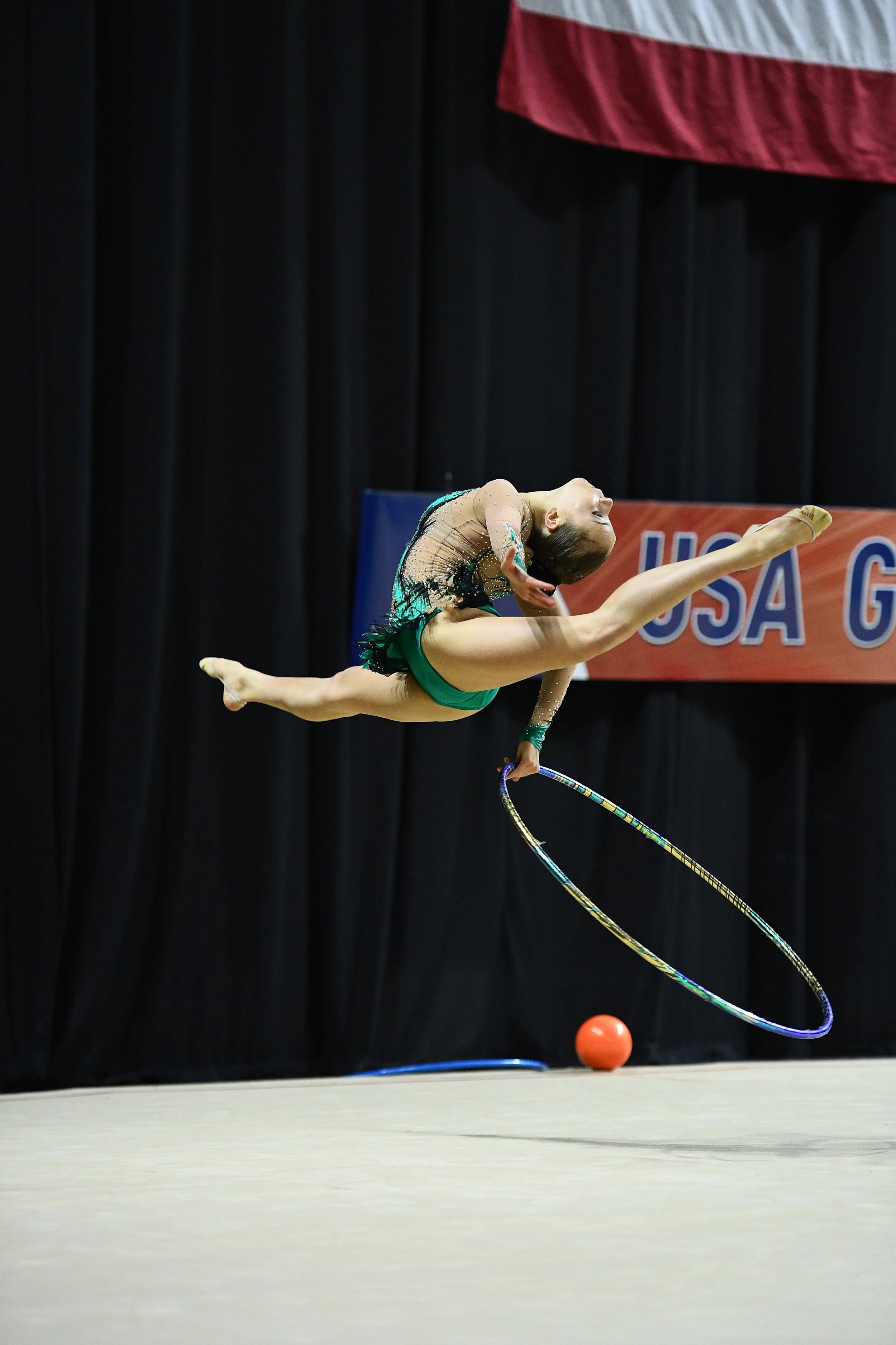 A young female gymnast performing a routine with a hoop on a stage with a black curtain background. An American flag and a sign reading "USA G" are visible behind her.