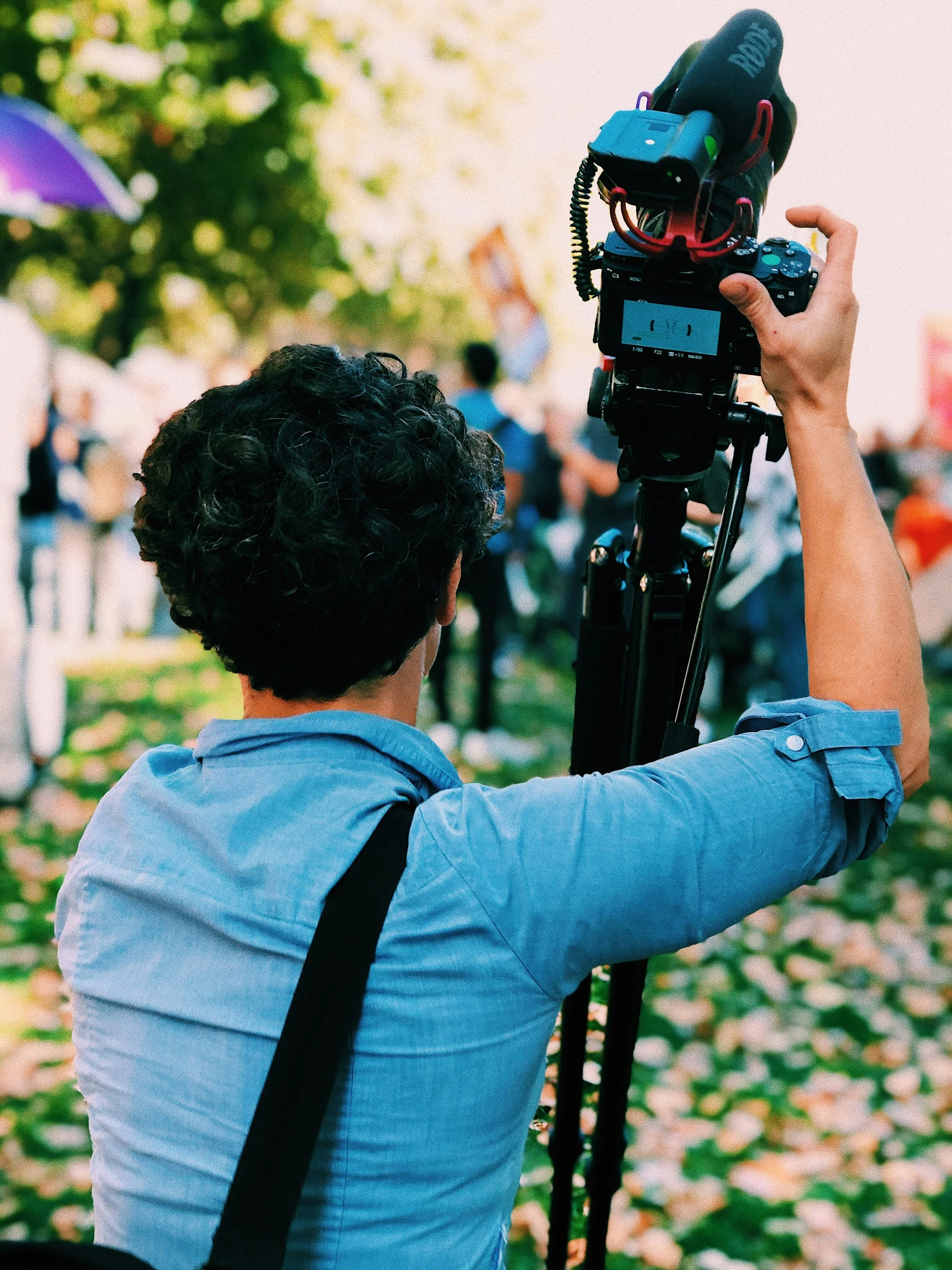 Photographer operating a DSLR camera on a tripod at an outdoor event, with a crowd and trees in the background.