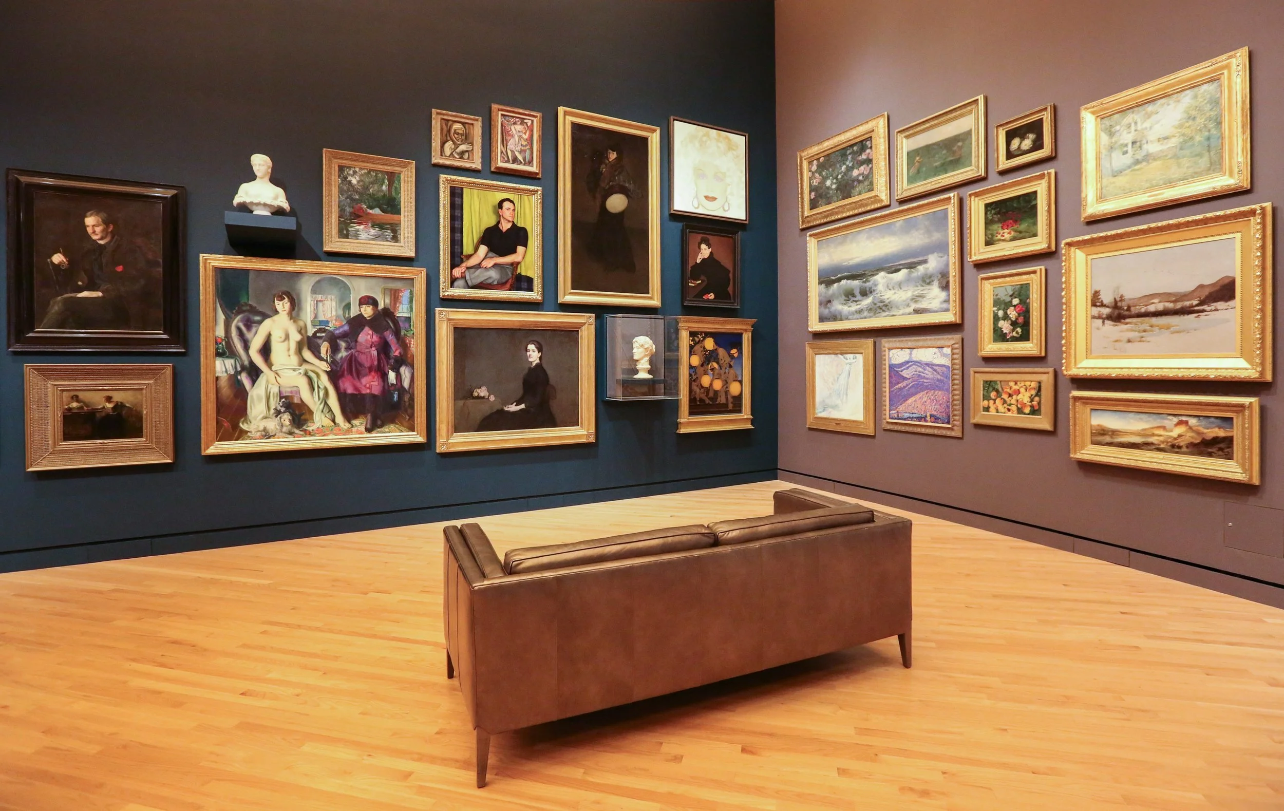 An art gallery with a variety of framed paintings on dark blue and gray walls, a brown leather couch centered on a wooden floor, and several small sculptures and decorative items displayed among the paintings.