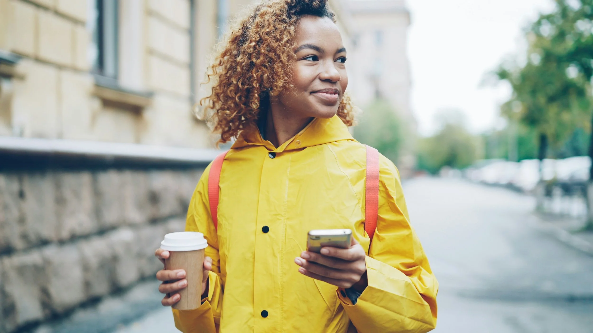 A young woman with curly hair wearing a yellow rain jacket holding a takeaway coffee cup and a smartphone, standing outdoors on a city street during daytime.