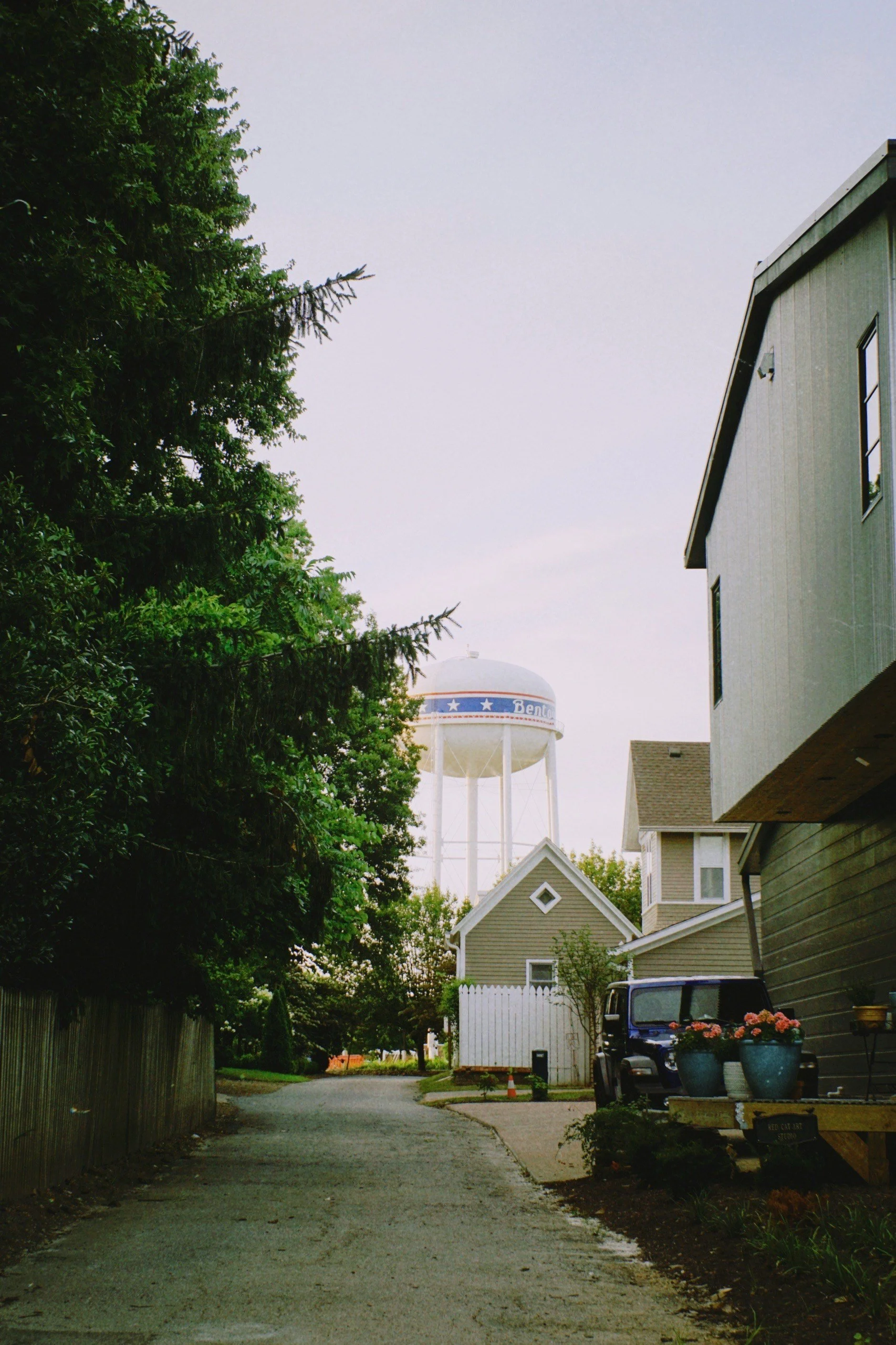 Residential alley view with houses, a black vehicle, potted flowers, trees, and a large water tower in the background.