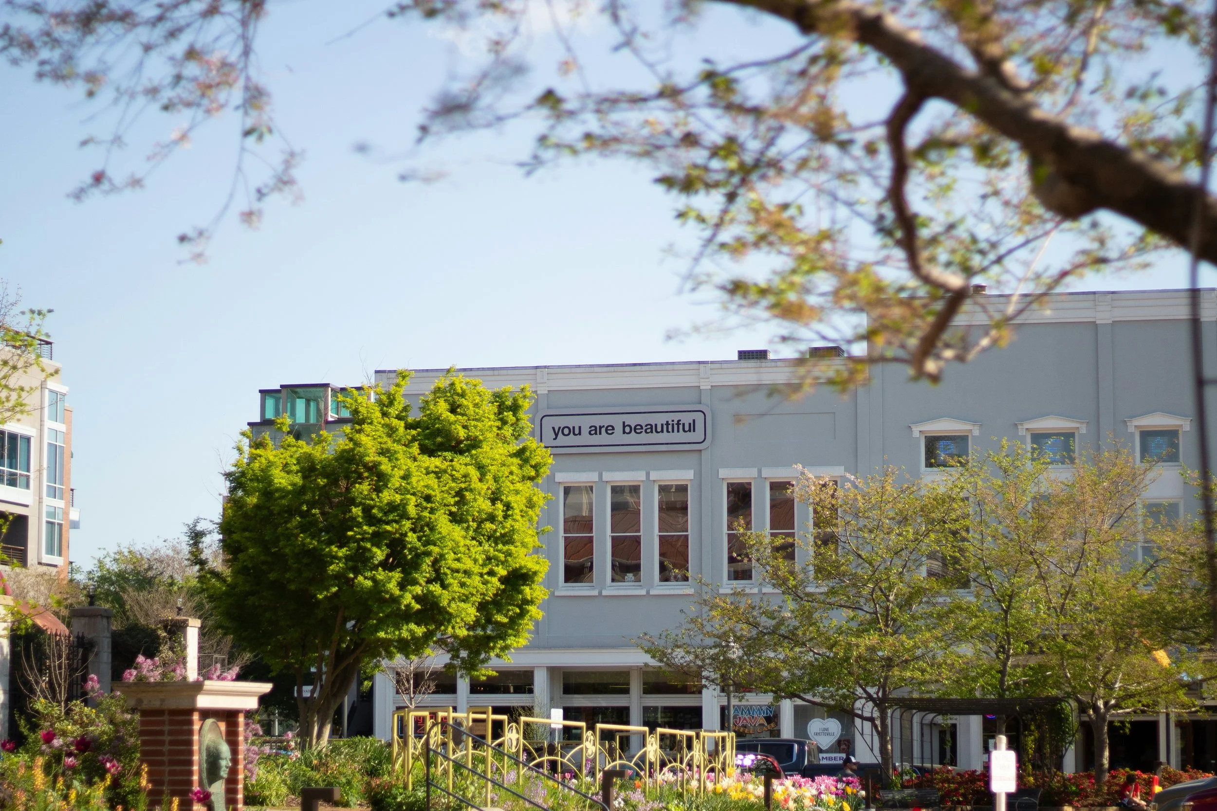 A white building with a sign that says "you are beautiful," surrounded by trees and colorful flowers in a park or garden setting.