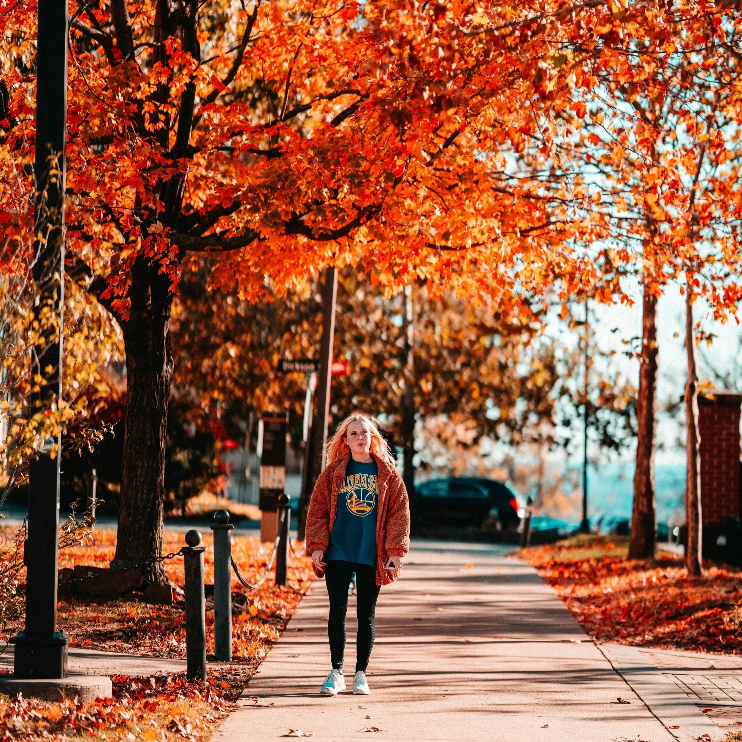 A young woman walking on a sidewalk lined with orange and red autumn leaves, under trees with colorful fall foliage, during daytime.