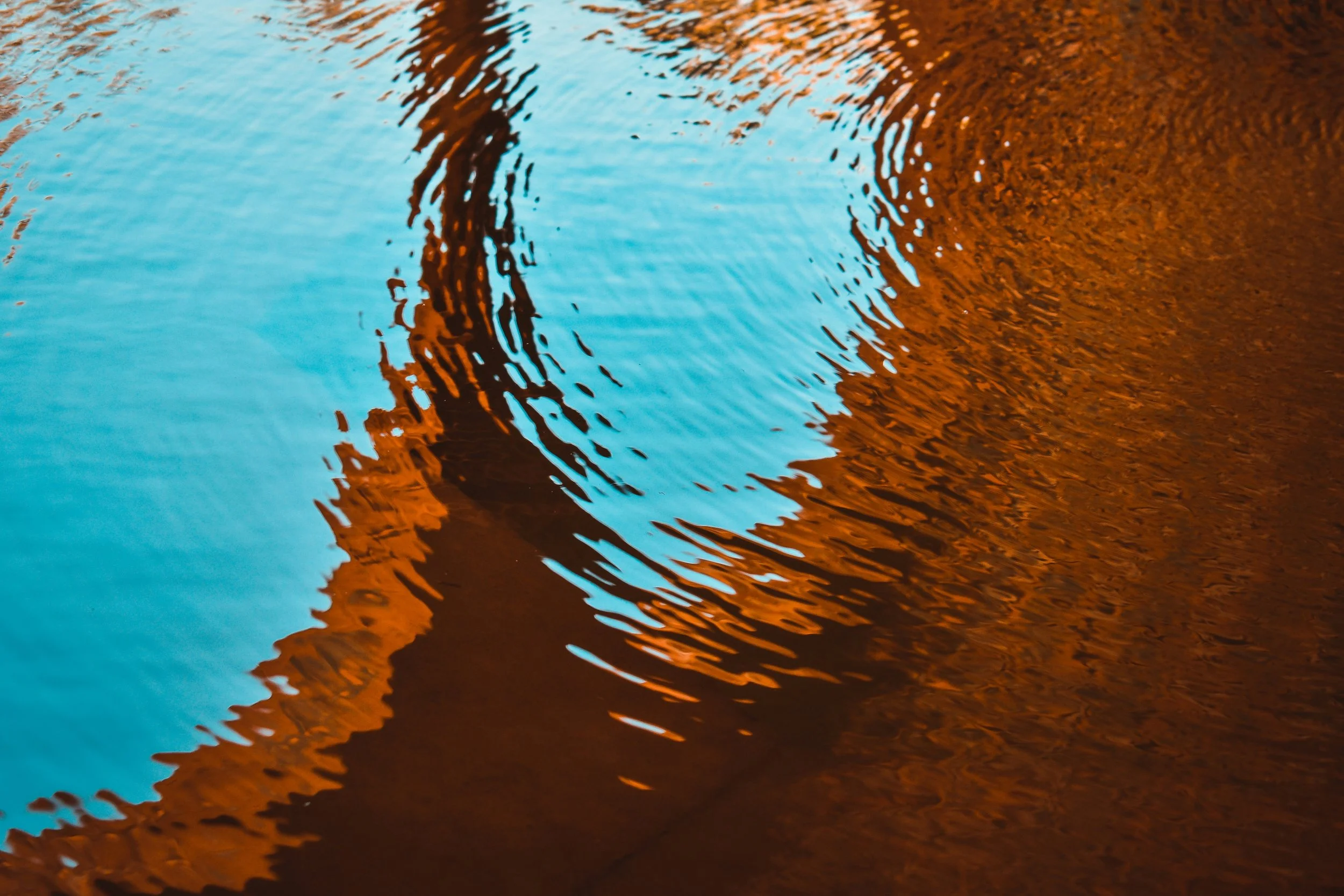 Reflections of a bridge and surrounding trees in a body of water, with portions of the sky visible, creating a mixture of blue and reddish-brown hues.