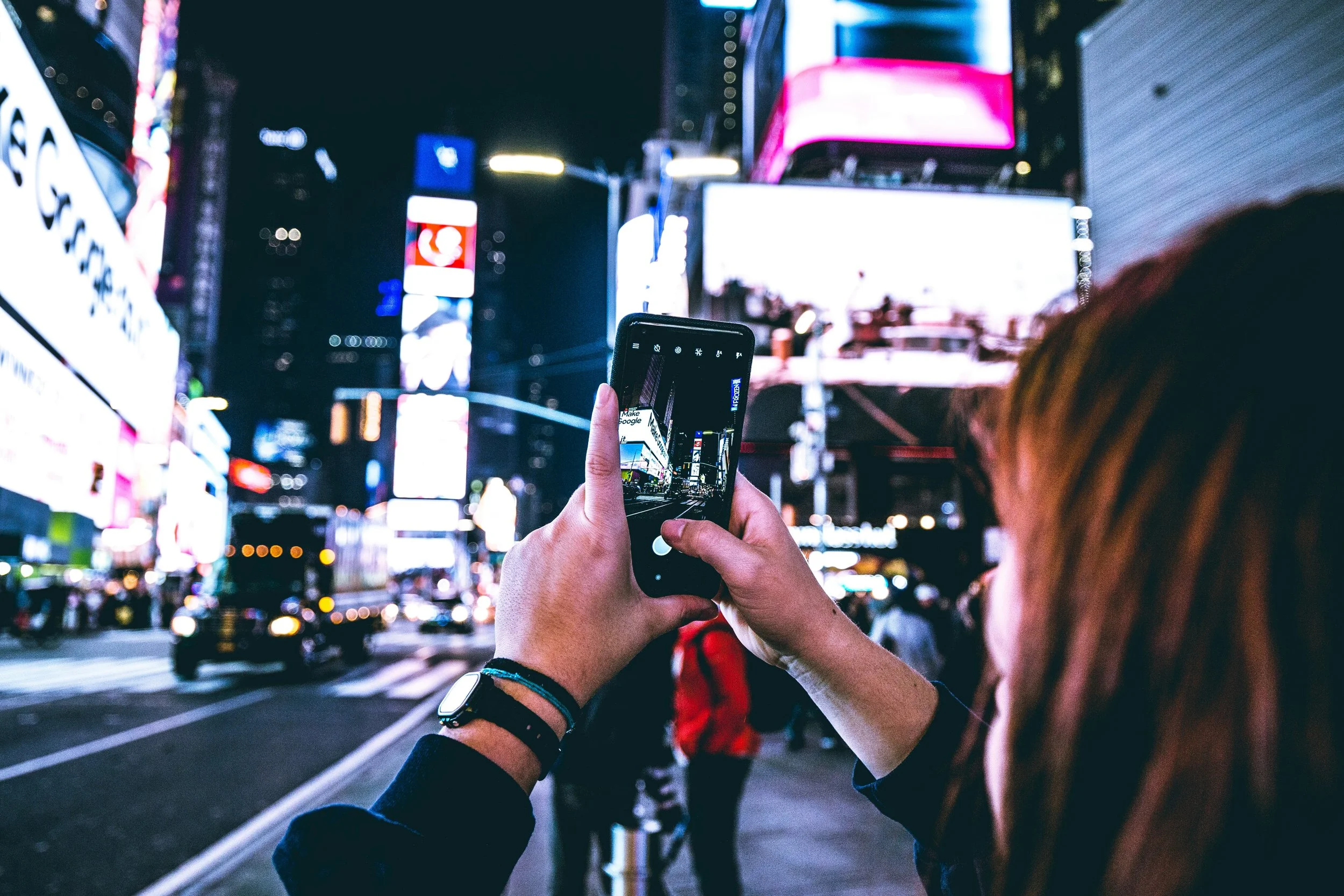 Person taking a photo with a smartphone on a busy city street at night, illuminated billboards and city lights in the background.