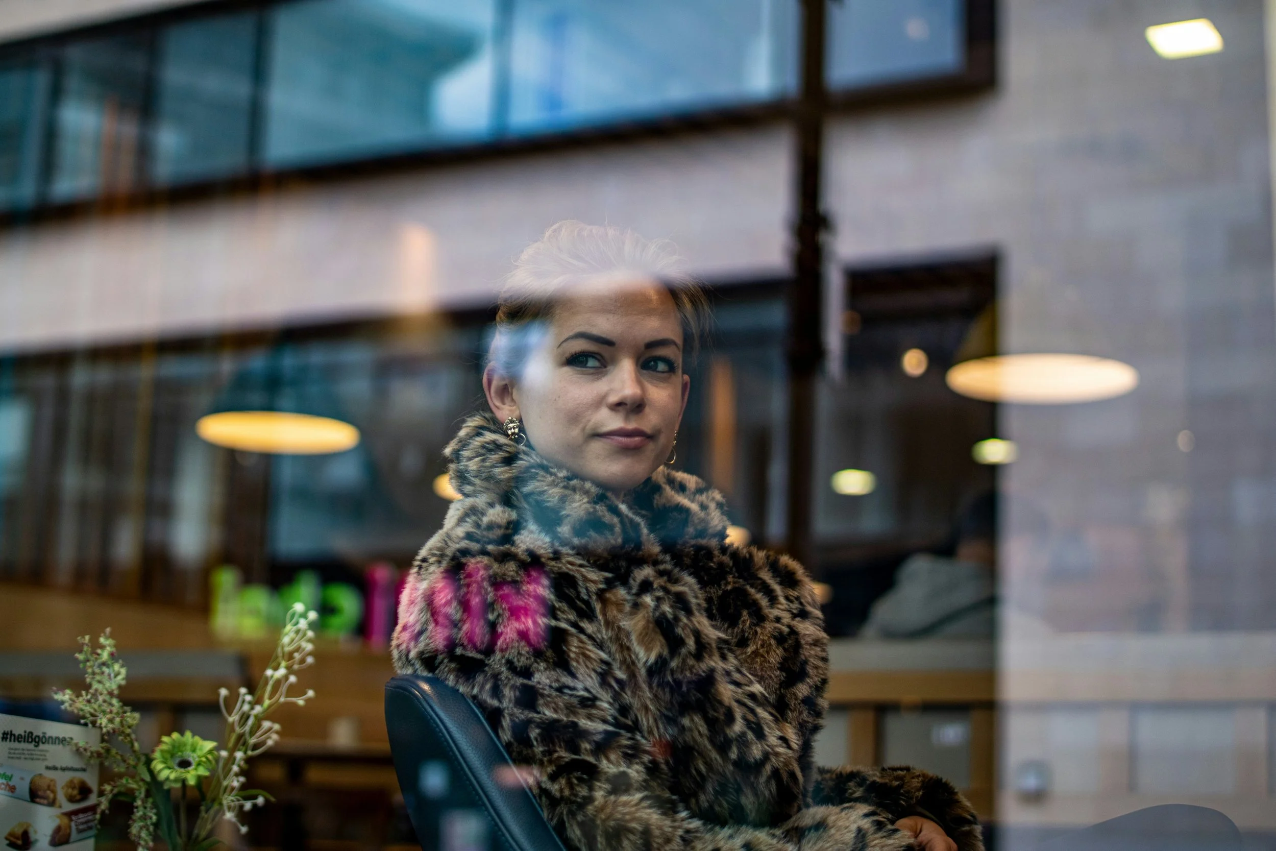A woman in a leopard-print fur coat sitting inside a cafe, looking out the window with a slight smile. The reflection of the interior lights and decor can be seen on the glass.