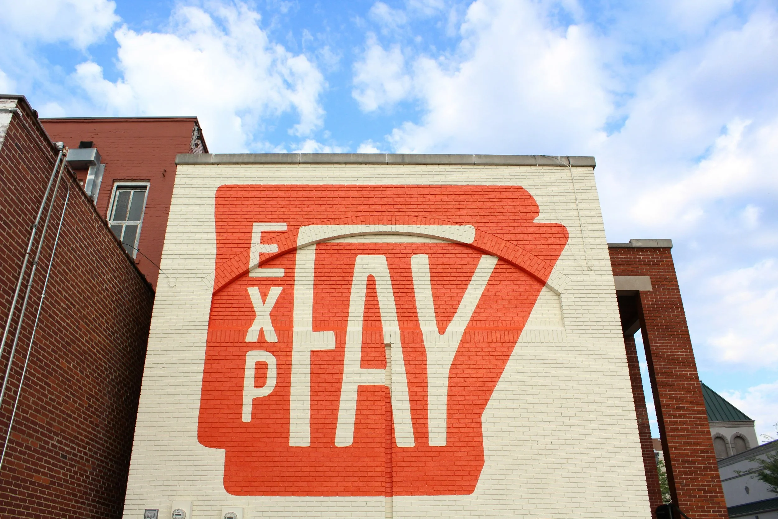 Large mural on a brick building with the words 'Explore, Play' in bold, stylized letters, designed to resemble a popsicle or ice cream treat, against a bright blue sky with some clouds.
