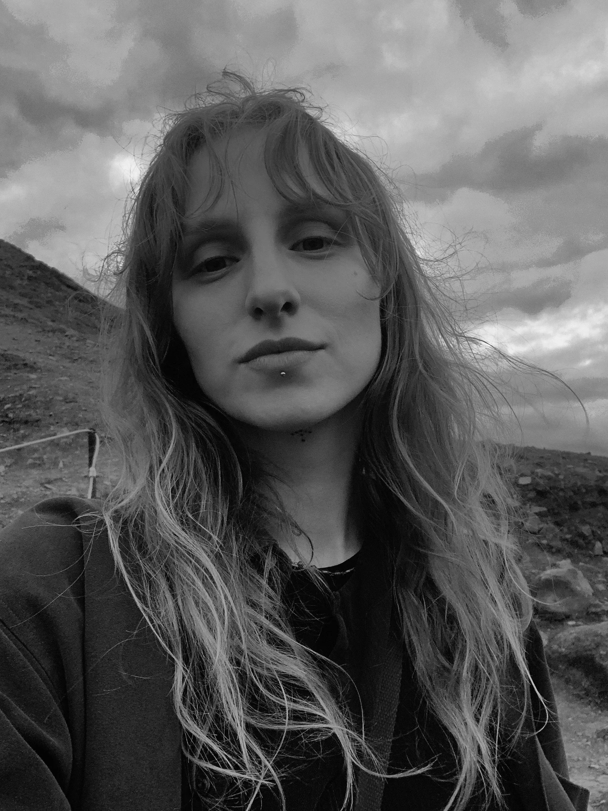A young woman with long, wavy hair and a lip piercing taking a selfie outdoors on a cloudy day, with a hillside and cloudy sky in the background.