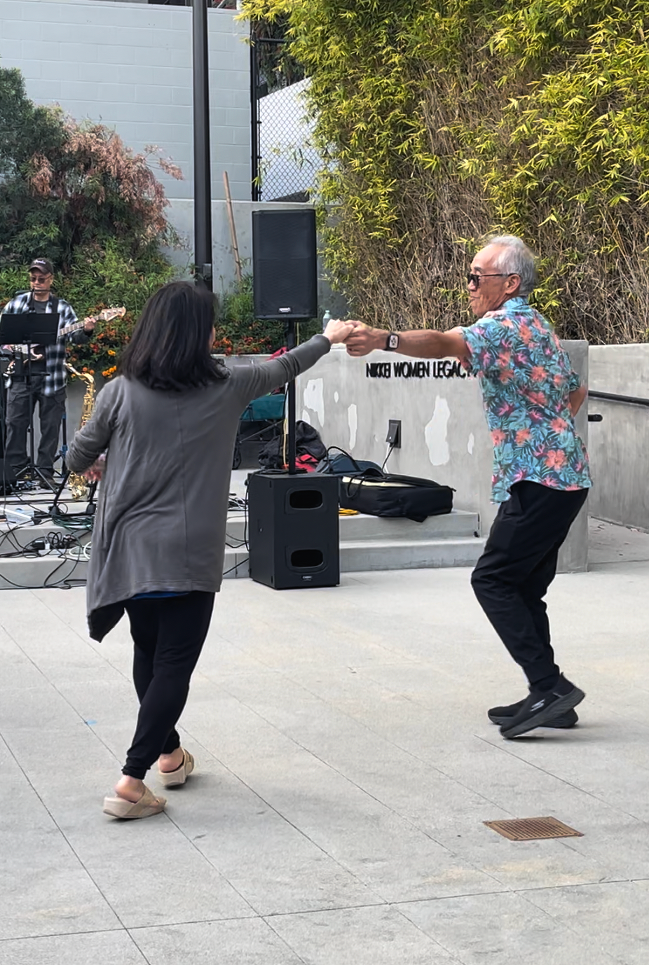 An elderly man in a colorful Hawaiian shirt and black pants is dancing with a woman in a gray jacket and black pants at an outdoor event, with a musician playing guitar and saxophone performing in the background.