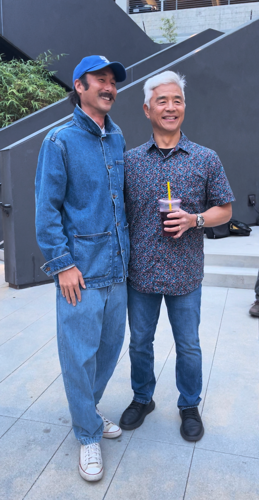 Two men smiling and standing together outdoors. One is wearing a denim jacket, jeans, and a blue cap. The other is wearing a floral short-sleeve shirt, jeans, and holding a drink with a yellow straw. They appear happy and are posing for a photo.