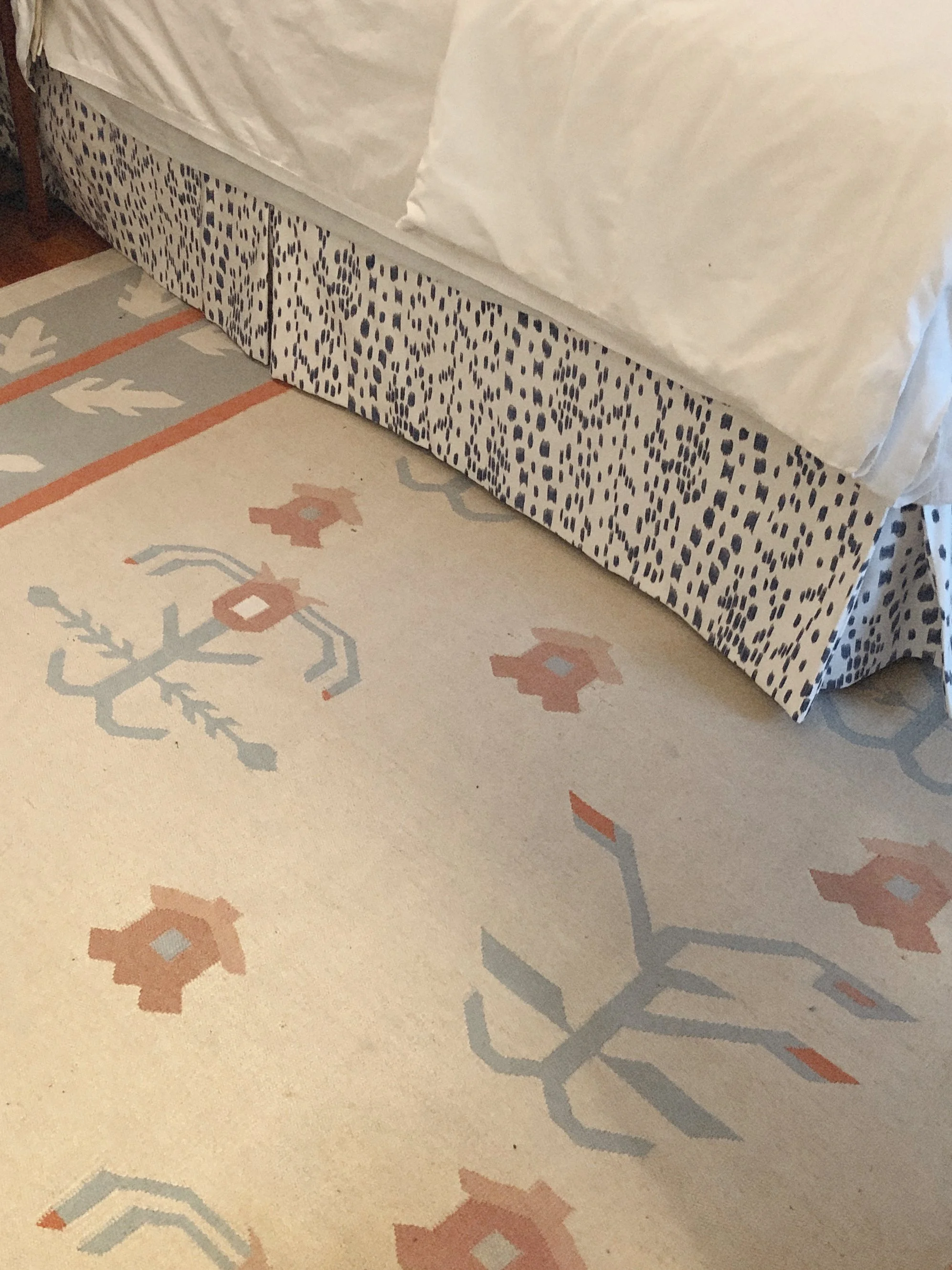 Close-up of a bed with a patterned bed skirt and white bedding, and a cream-colored rug with geometric and floral designs in peach, light blue, and gray.