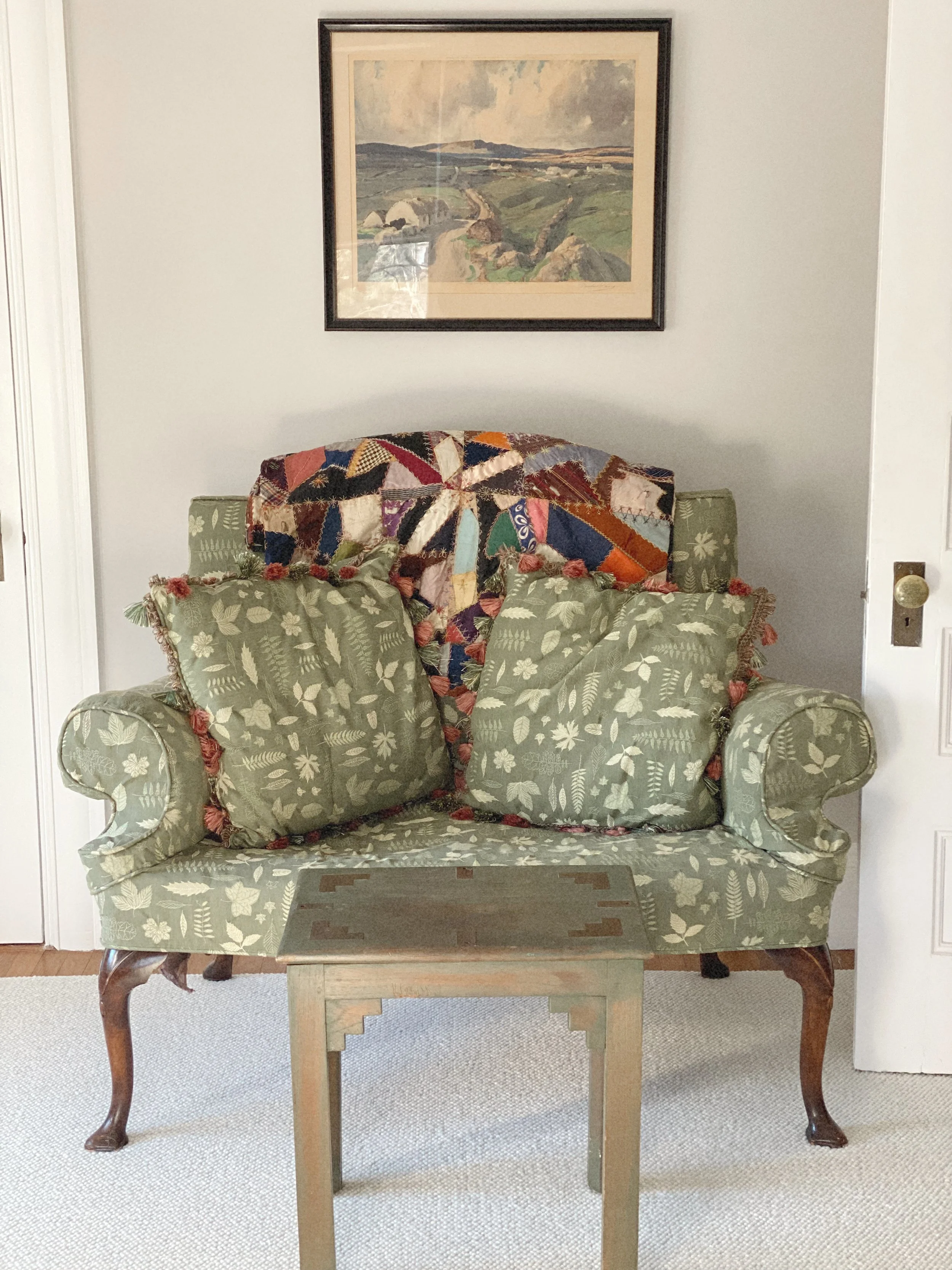 A vintage green floral upholstered loveseat with two matching pillows, a multicolored patchwork quilt draped over the back, a small wooden coffee table in front, and a landscape painting hanging above on a white wall.