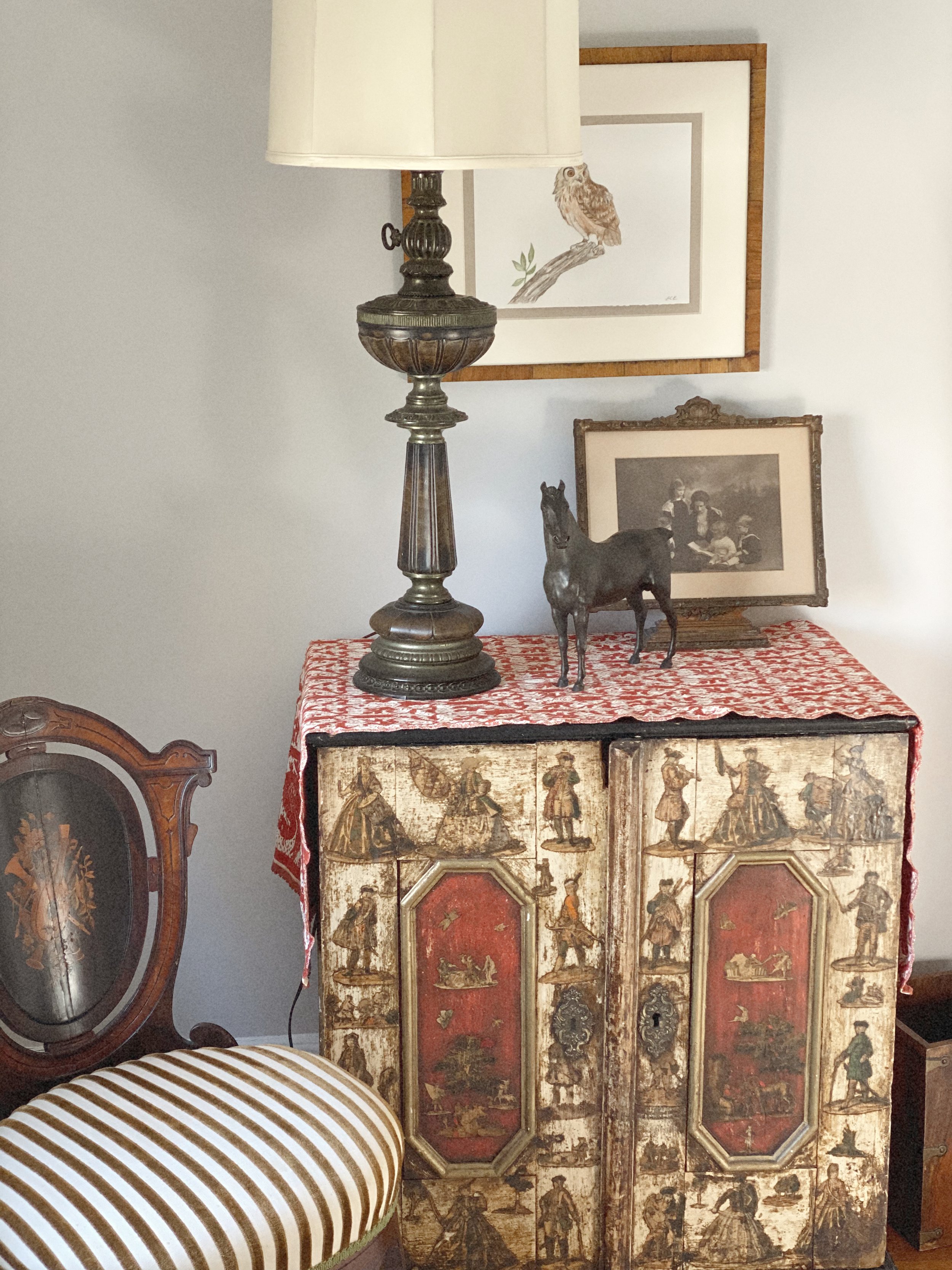 An antique wooden cabinet with painted figures, topped with a red and white cloth, a vintage table lamp, a small metal horse sculpture, and framed artwork, including one of an owl on a branch and another of three women.