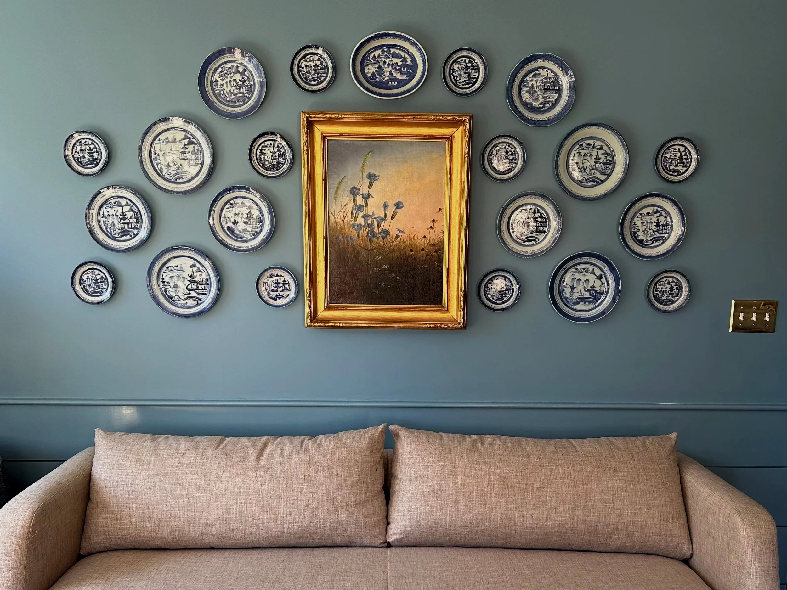 Living room wall decorated with a central framed landscape painting and surrounding blue and white decorative plates, with a beige fabric sofa at the bottom.