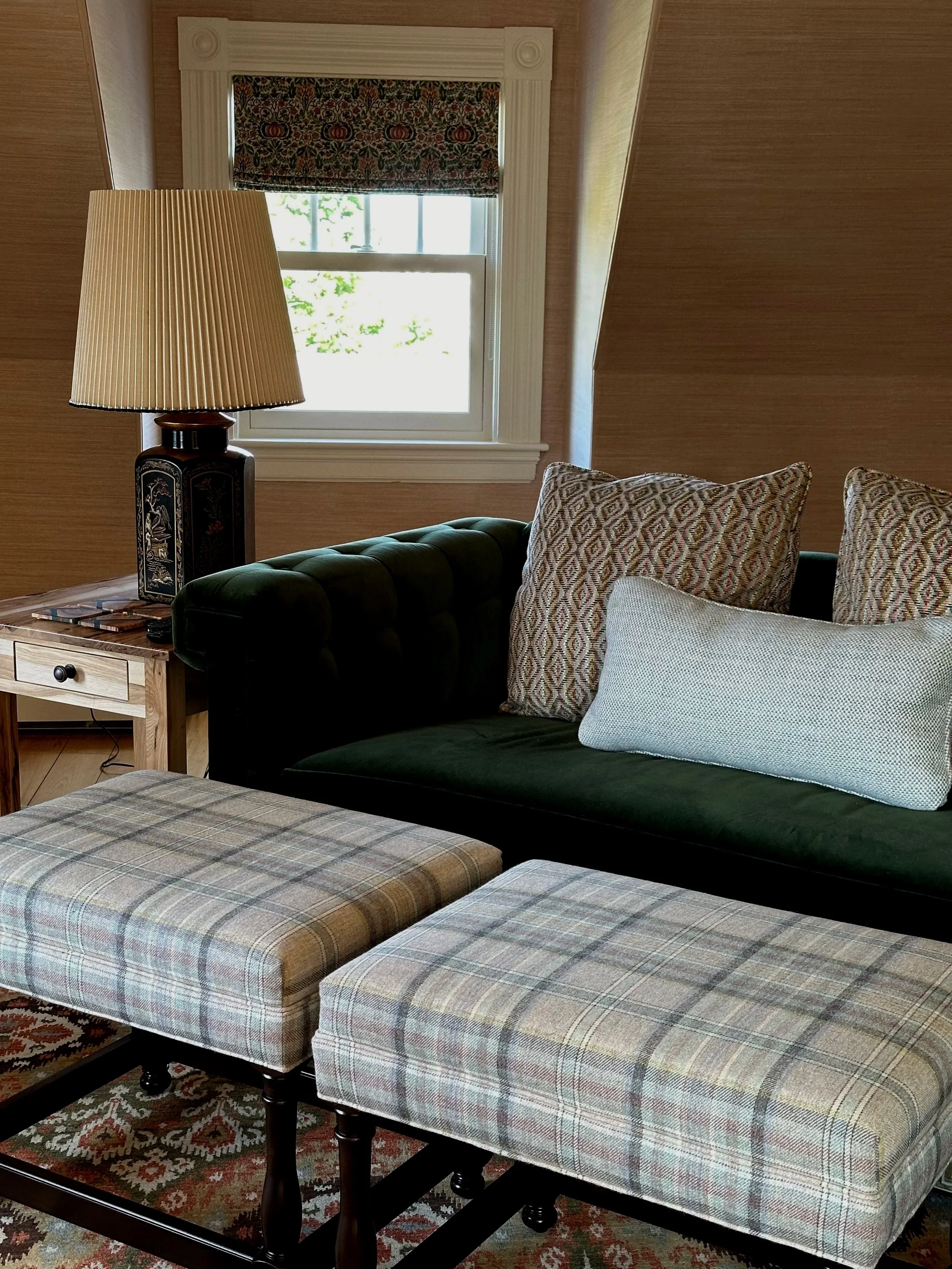 A cozy living room corner with a green velvet sofa, decorative pillows, a wooden side table with a lamp, a window with floral valance, and plaid upholstered stools.