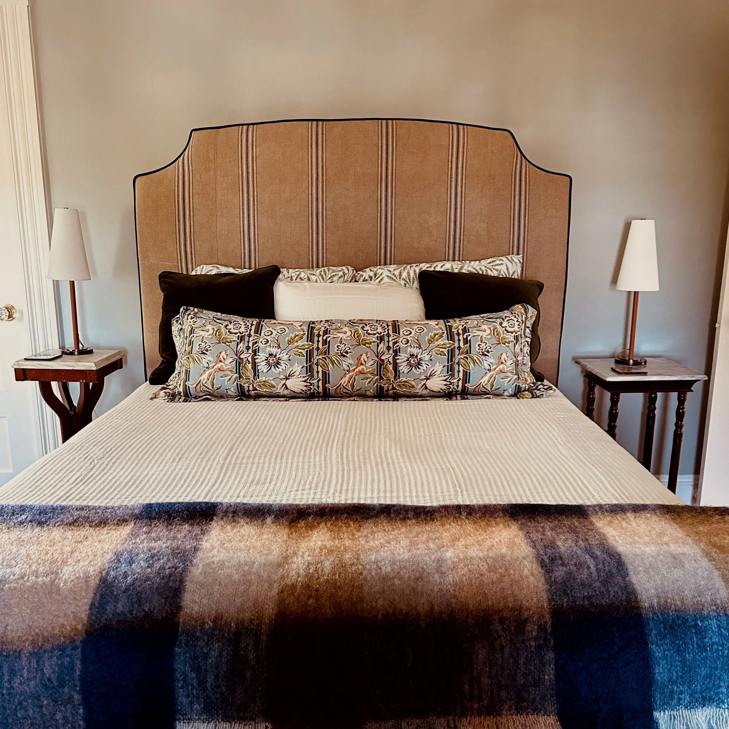 A neatly made bed with a beige pagoda-style headboard, decorative pillows including a long floral pillow and two black pillows, a beige bedspread, and a multi-colored plaid blanket at the foot of the bed. There are two wooden nightstands with white l
