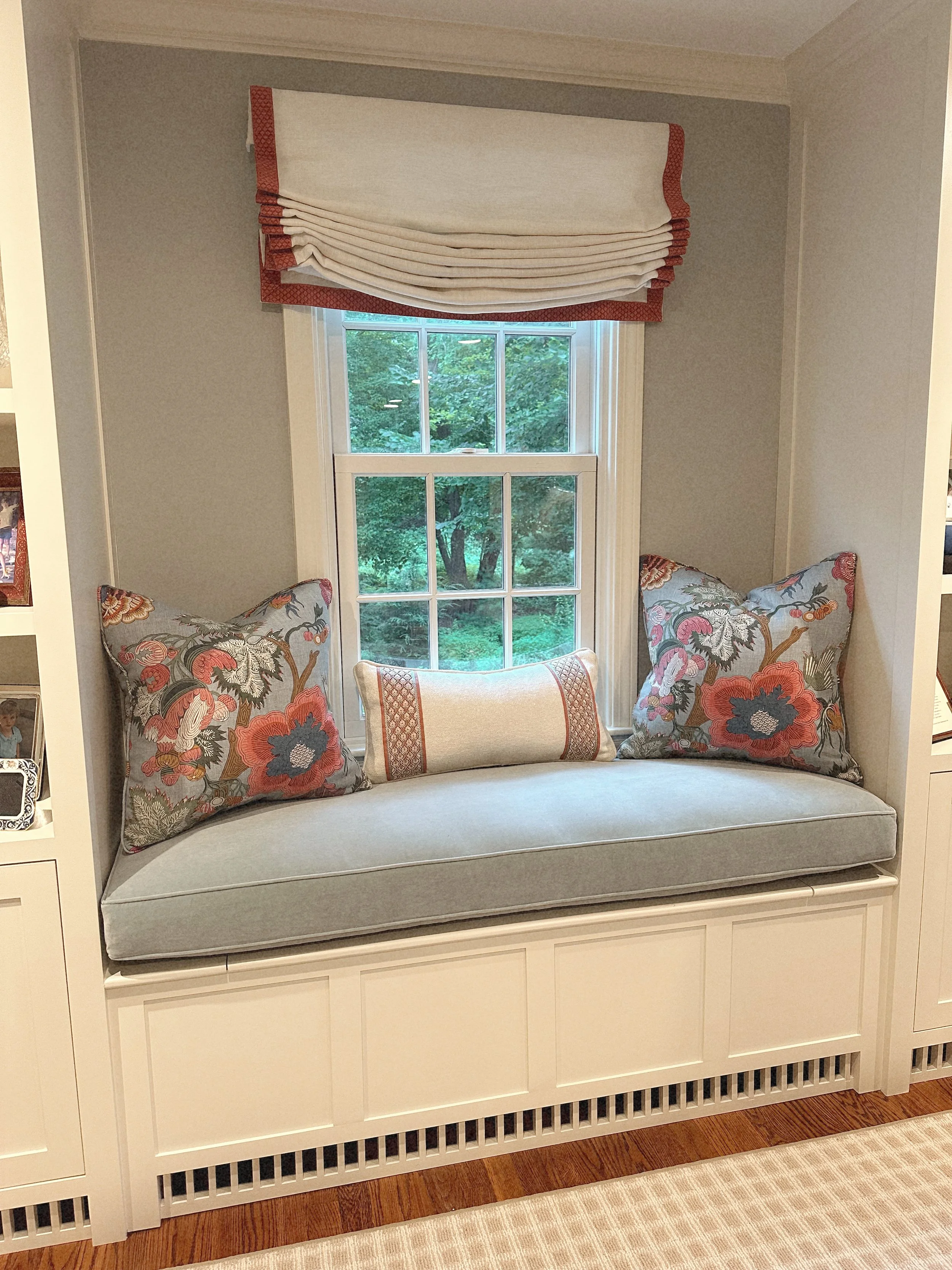 A window seat with a cushioned bench, decorative pillows, and a window with a white Roman shade valance, overlooking an outdoor green landscape.