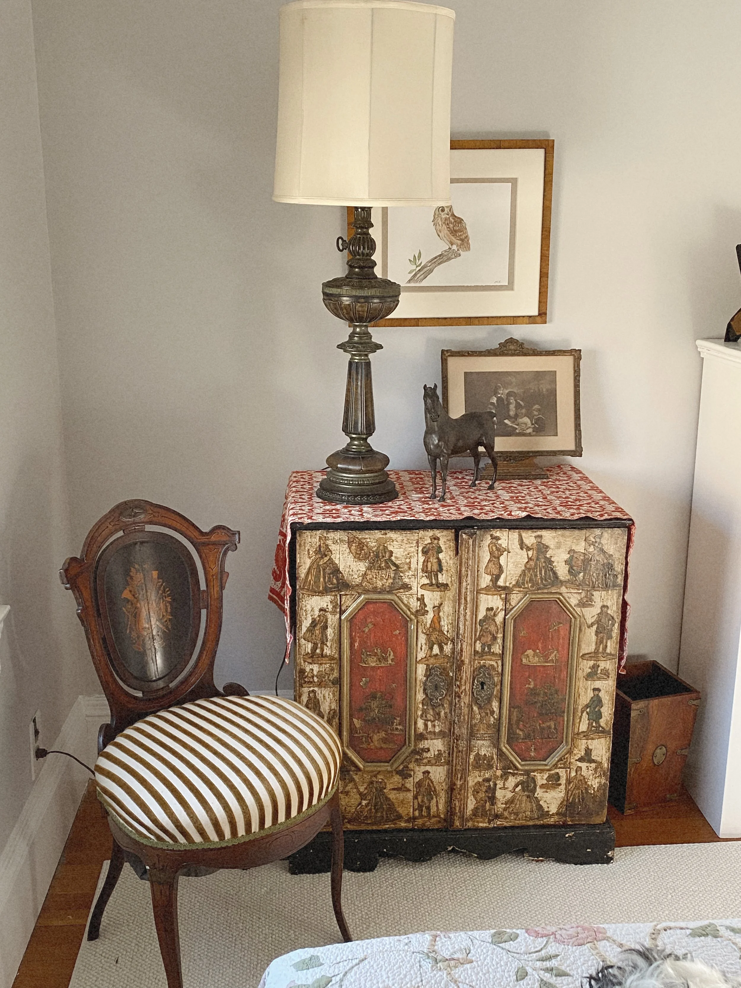 Decorative antique cabinet with a red and gold design, topped with a vintage tablecloth, shadows of framed art on the wall, a tall table lamp with a cream shade, a small horse figurine, a framed sketch of an owl, and a vintage wooden chair with strip