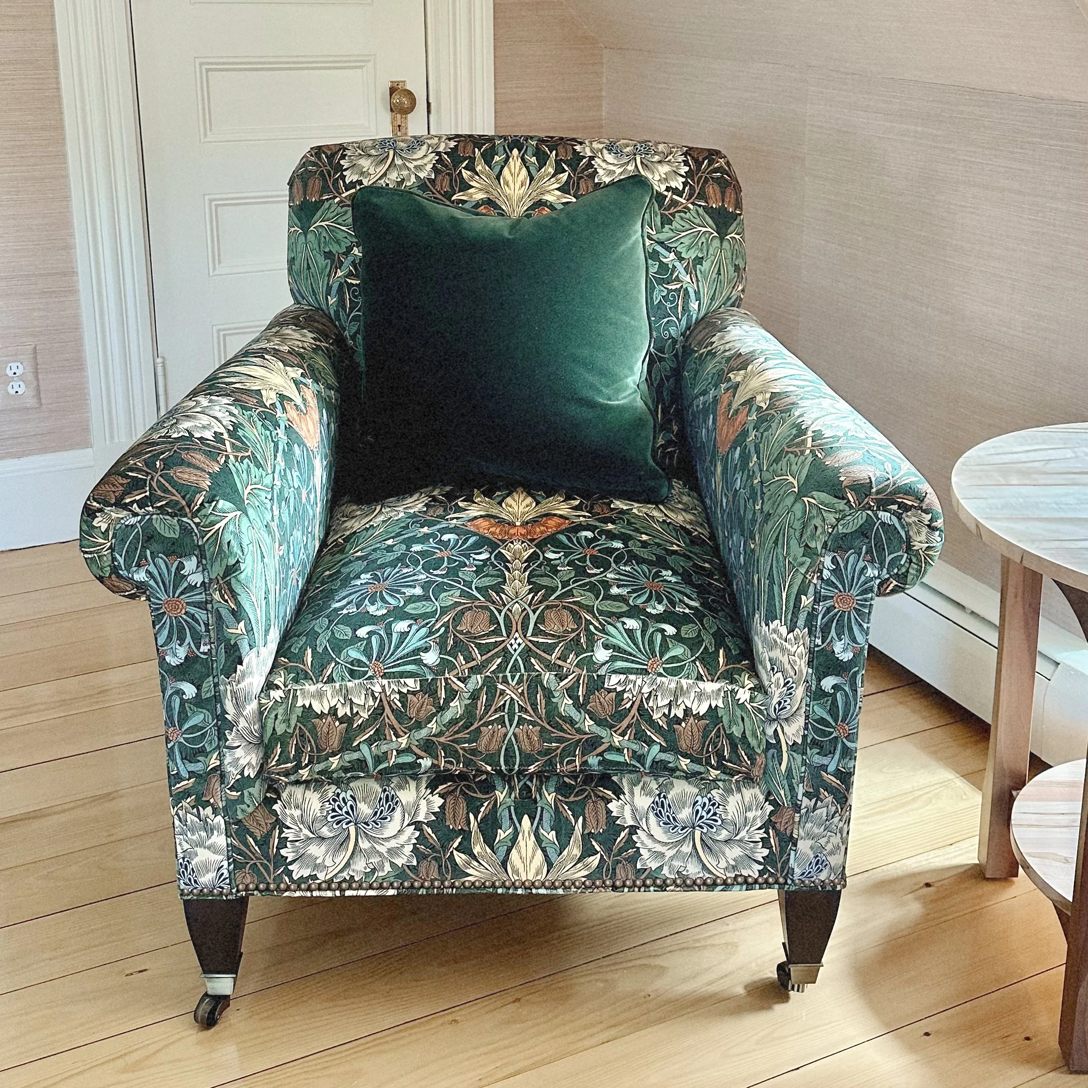A floral-patterned armchair with package floral fabric, dark wooden legs, and a dark green velvet cushion in a wood-floored room.