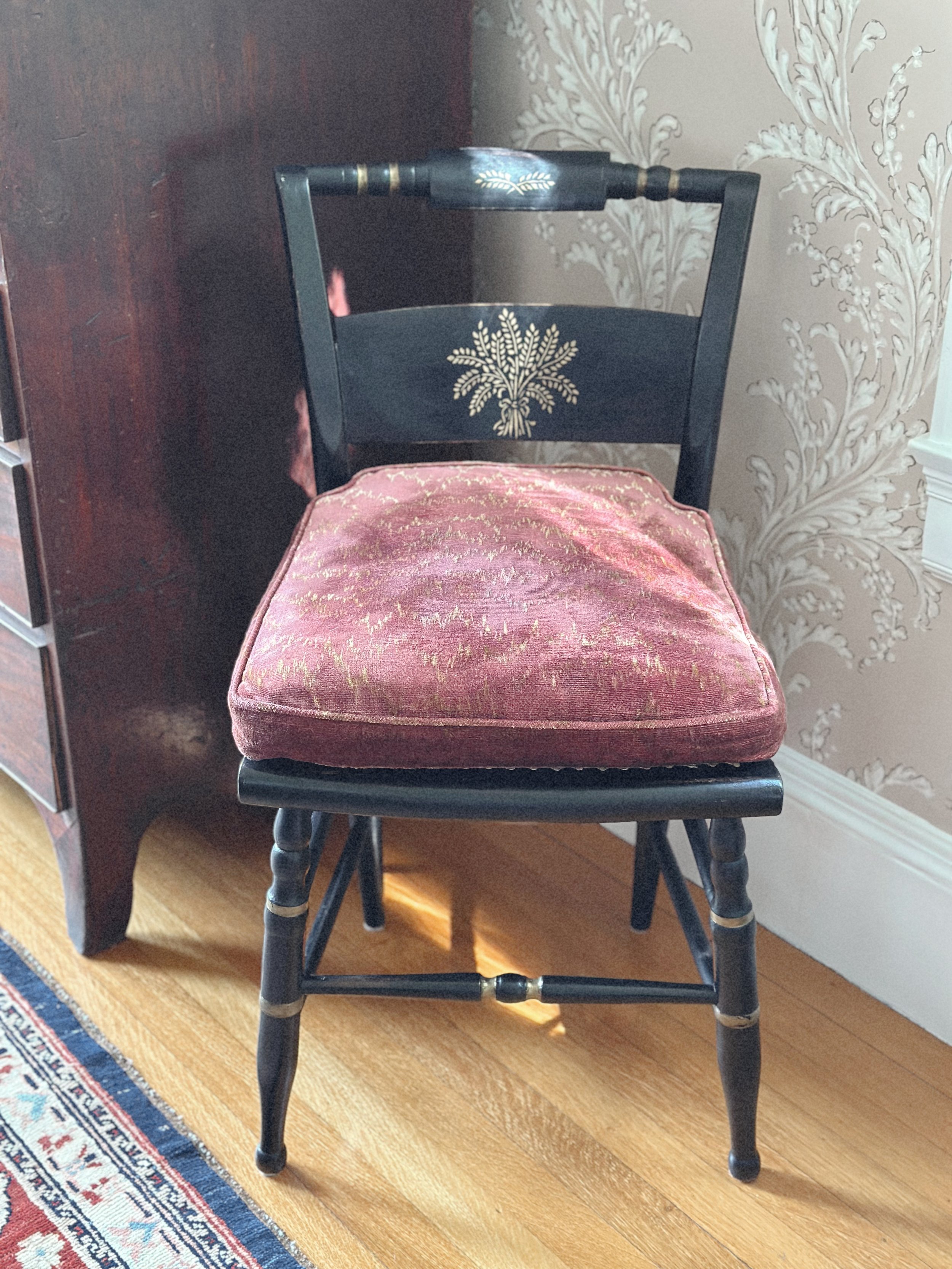 A vintage wooden chair with a pink velvet cushioned seat, gold accents, and a floral design on the backrest, placed on a hardwood floor next to a patterned wallpaper and a dark wooden piece of furniture.