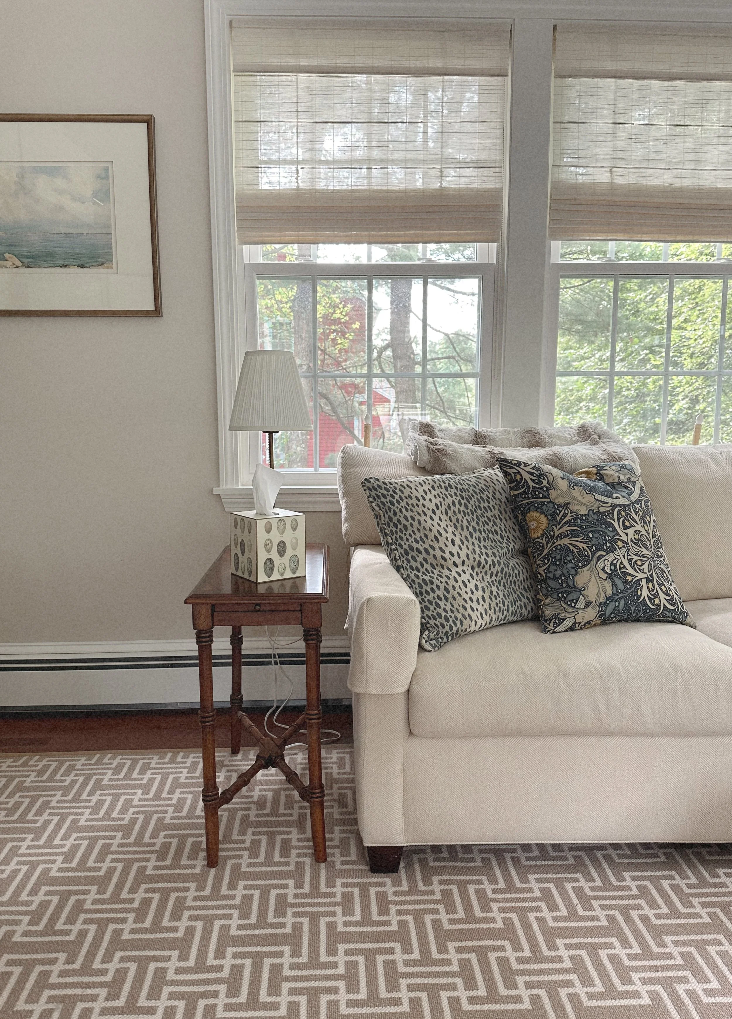 A cozy living room with cream-colored sofa, patterned cushions, a wooden side table with a lamp and tissue box, and large windows with blinds showing a view of trees outside.