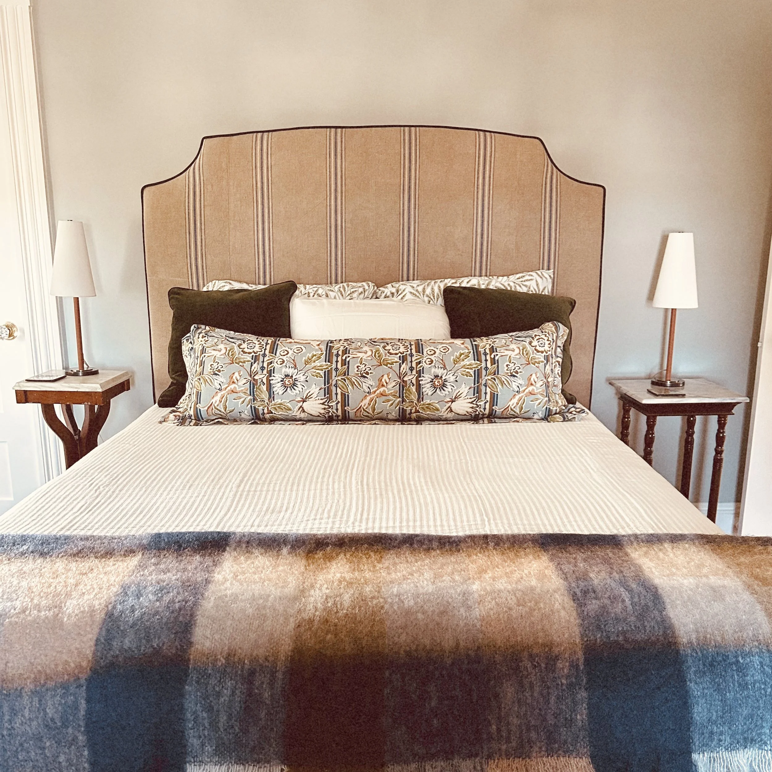 A neatly made bed with a beige and pink plaid blanket, a decorative floral pillow, and dark brown pillows, flanked by two wooden nightstands with white lamps, against a light-colored wall.