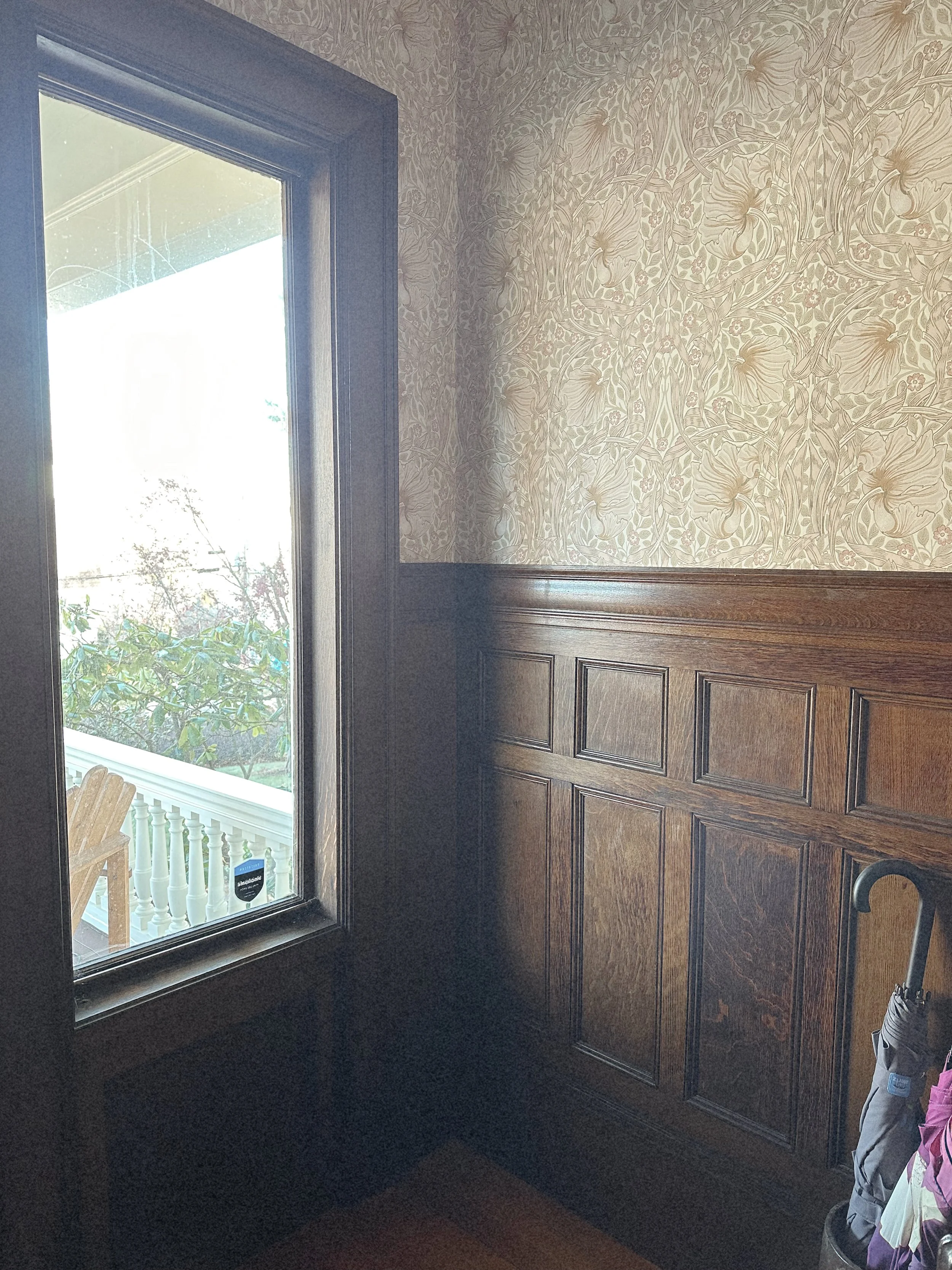 Interior corner of a room with wooden wainscoting on the walls, floral wallpaper above, and a large window showing an outdoor porch with a chair and greenery.