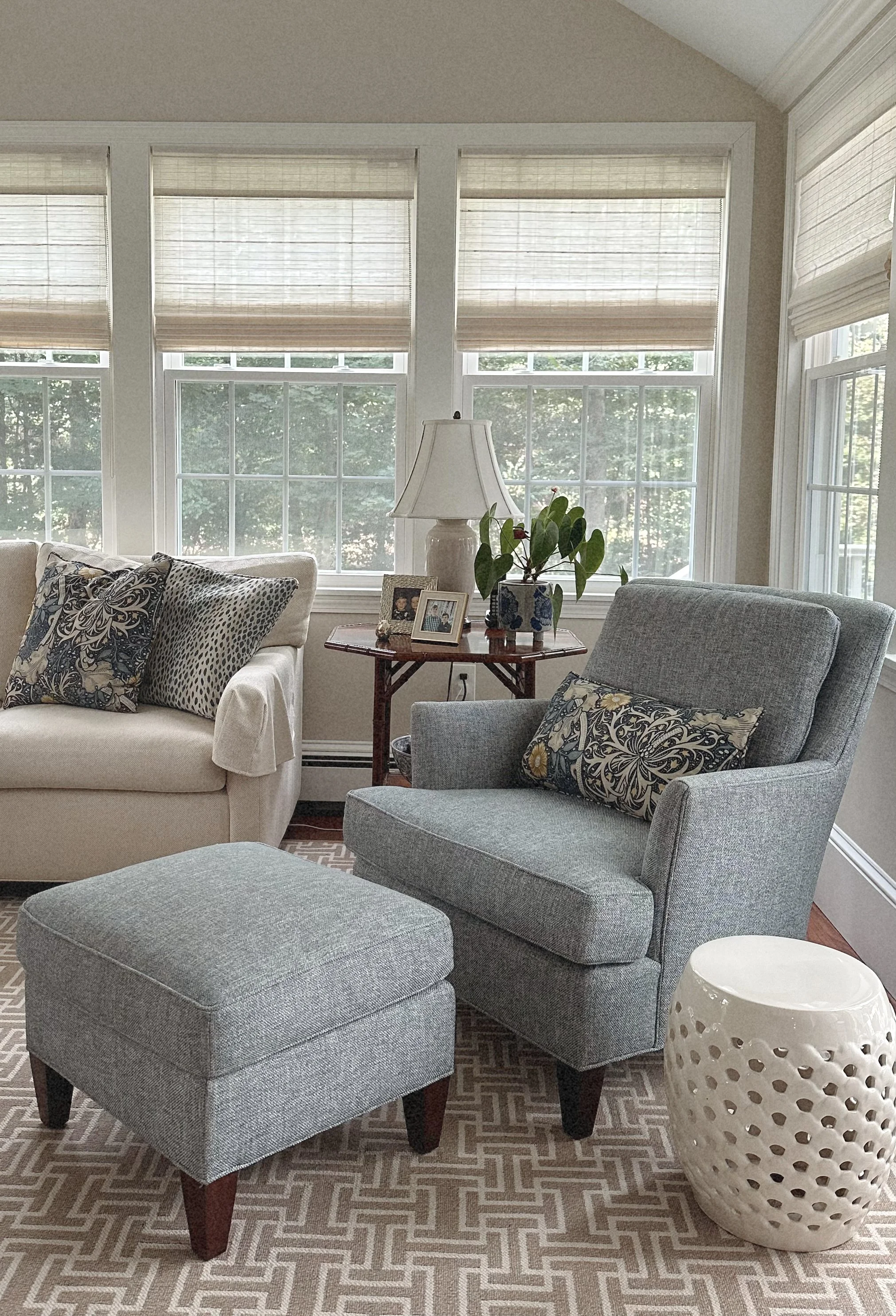 Living room with beige sofa, grey armchair with patterned pillows, small round wooden table with lamp and picture frames, potted plant on table, window with blinds, ceramic stool, and patterned rug.