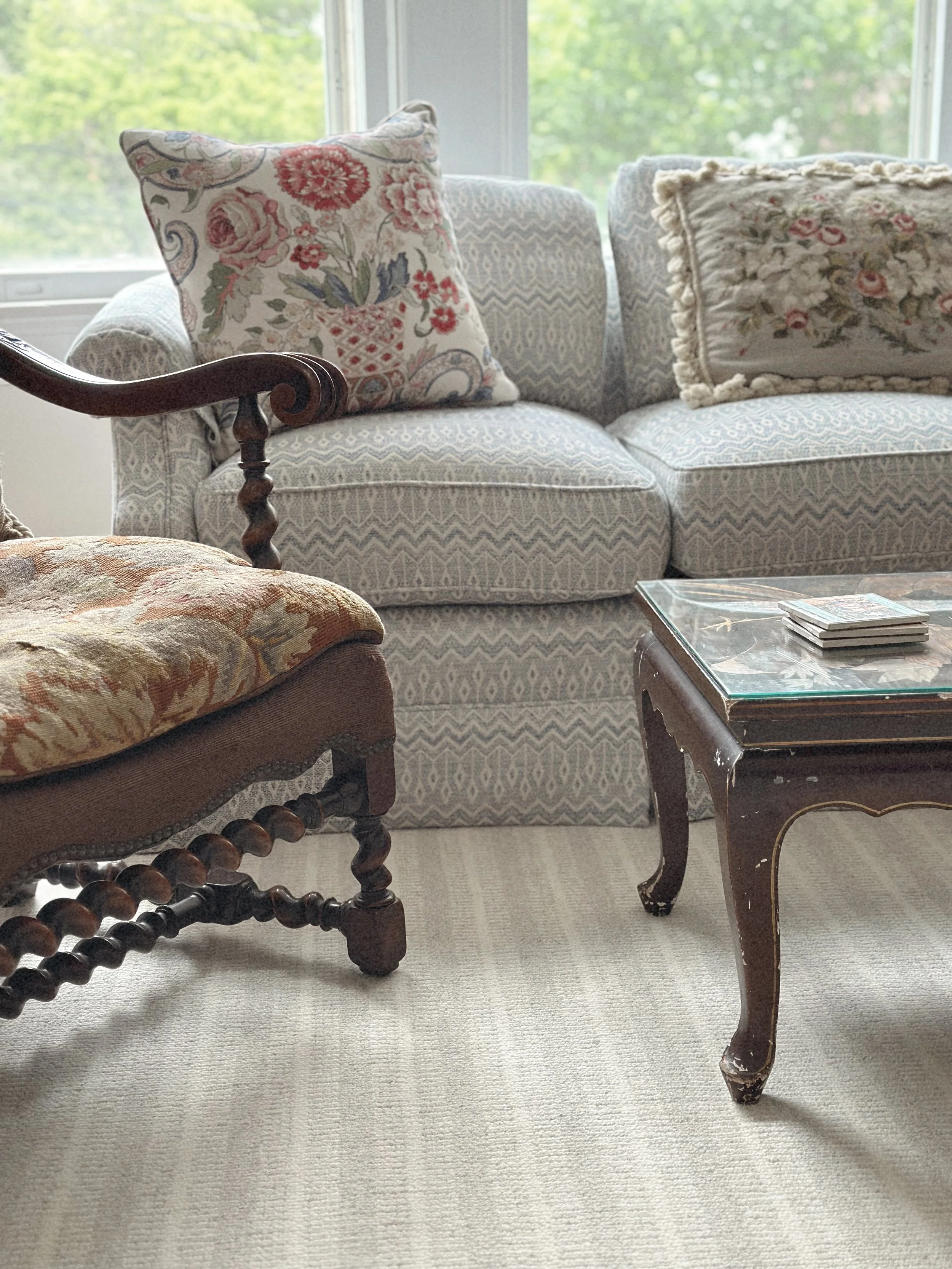 Living room with patterned sofa, decorative pillows, a vintage wooden armchair, a coffee table with magazines, and a large window showing green trees outside.