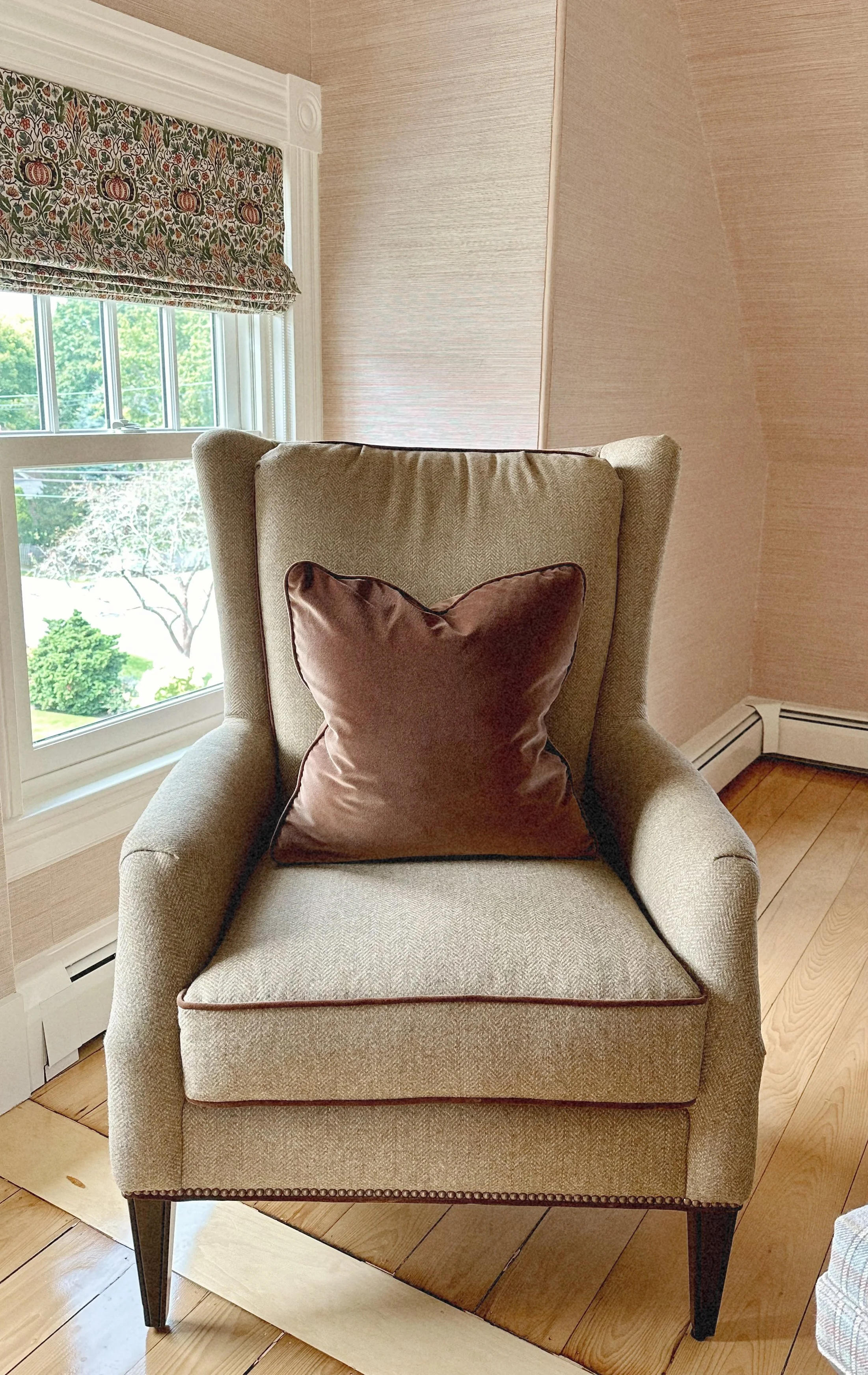 An upholstered armchair with a brown satin throw pillow, positioned near a window with floral patterned valance, on a wooden floor in a cozy room.