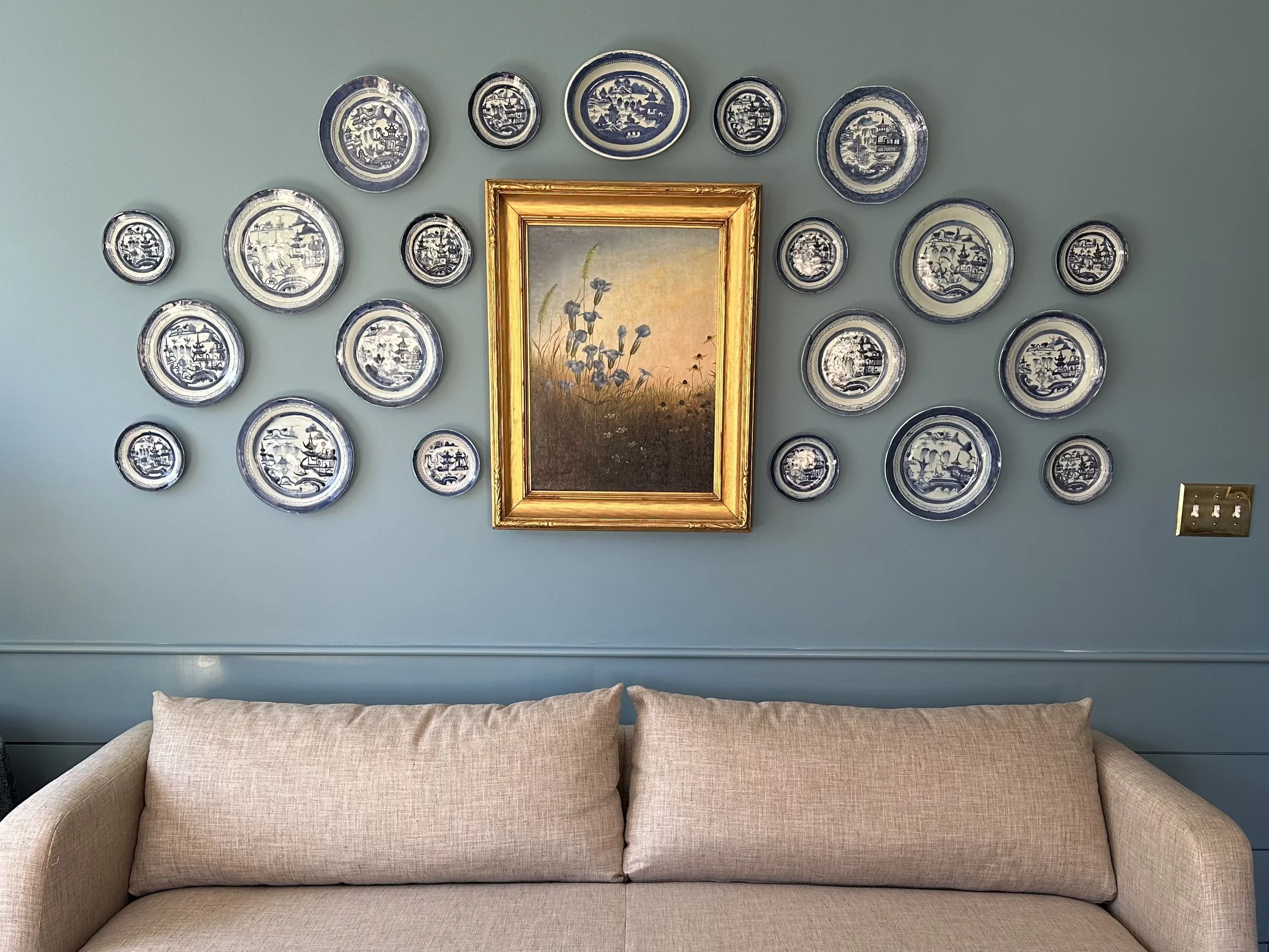 A beige sofa in front of a blue wall decorated with a collection of blue and white decorative plates and a framed painting of wildflowers.
