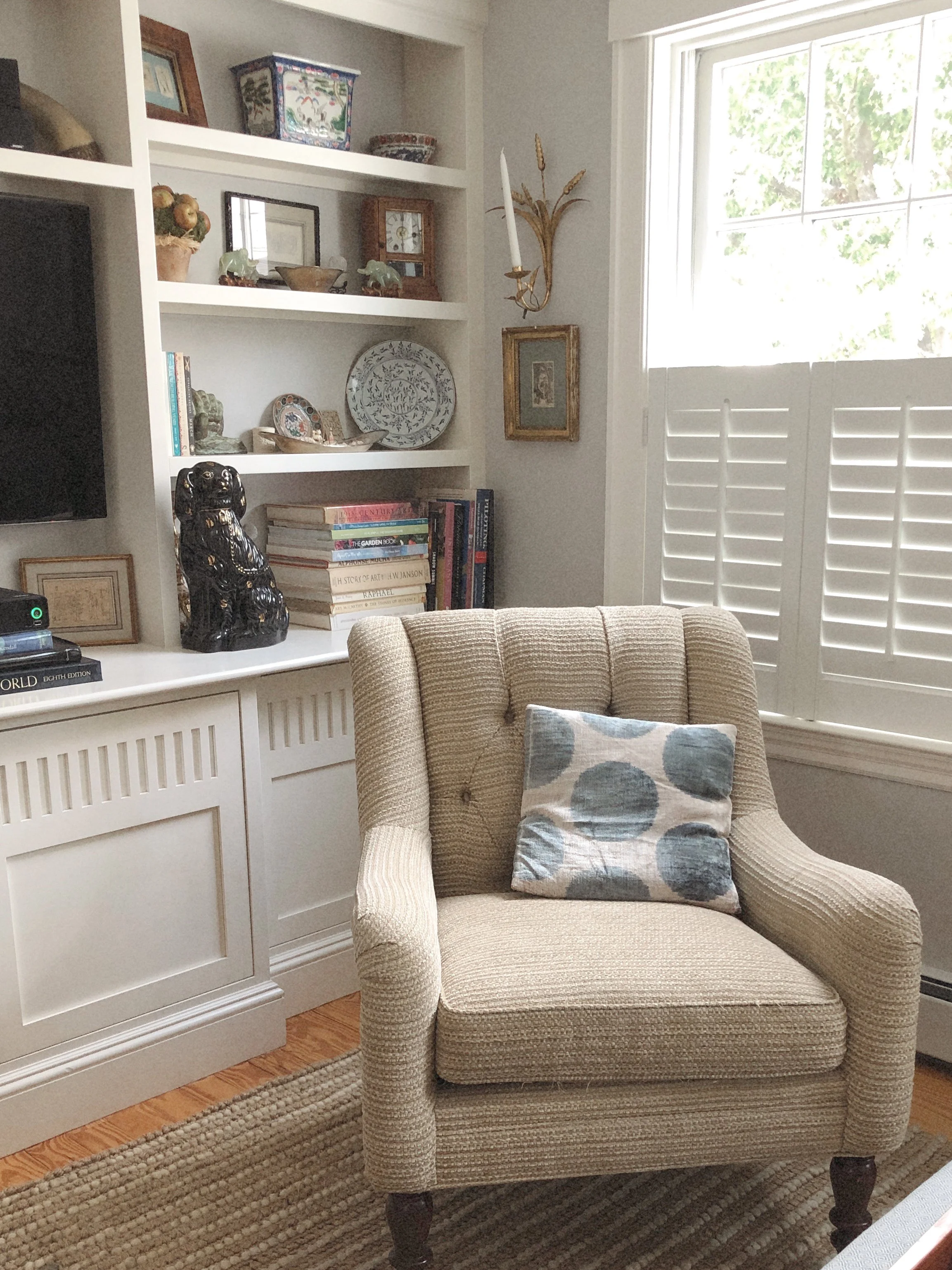 A cozy living room corner with a beige upholstered armchair that has a patterned cushion, a white built-in bookshelf filled with decorative items, books, and framed pictures, a window with white shutters letting in natural light, and a hardwood floor