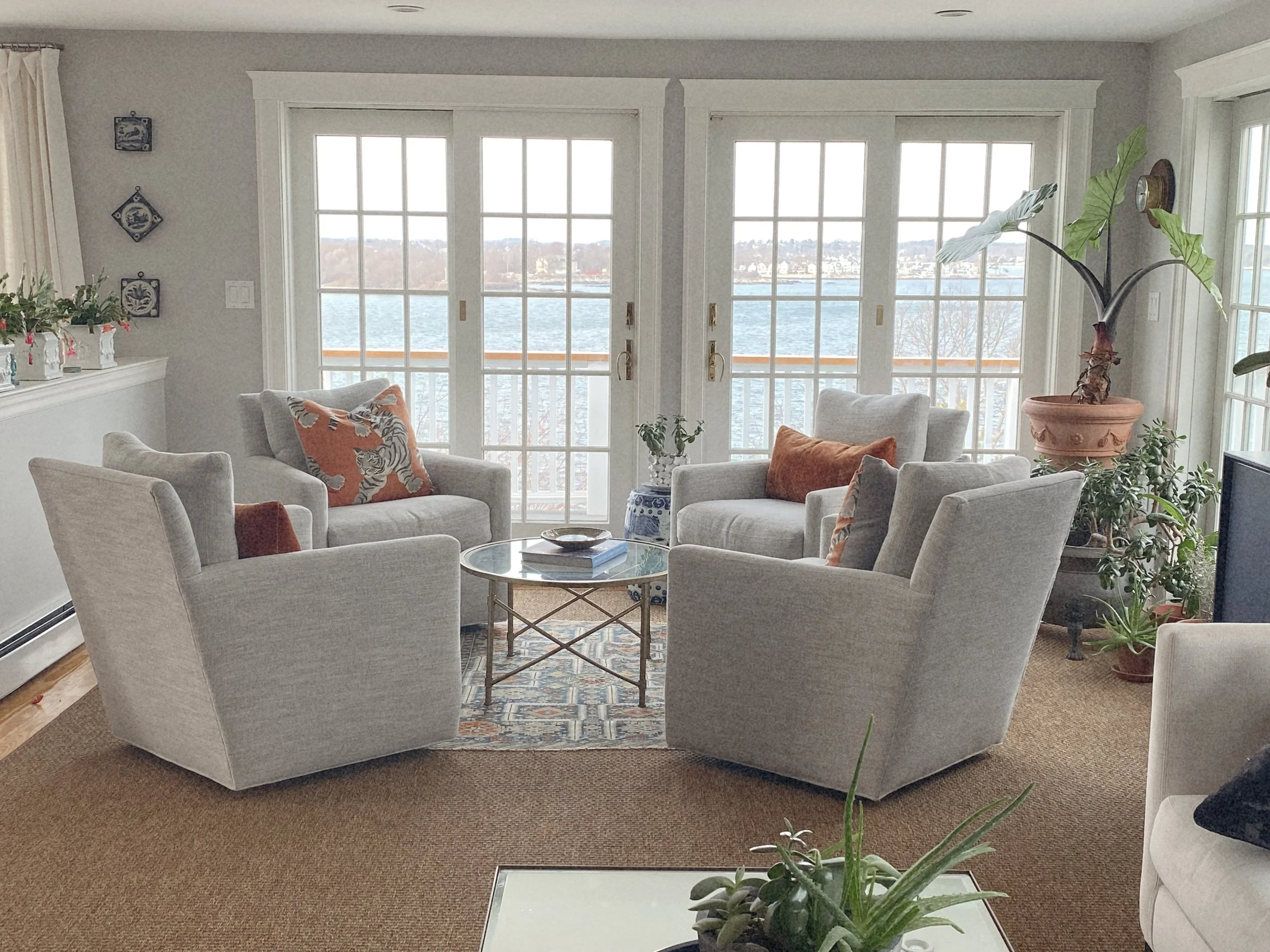 Living room with four light gray armchairs and a glass coffee table on a patterned rug, facing large windows showing a body of water and trees outside, with plants and decorative pillows.