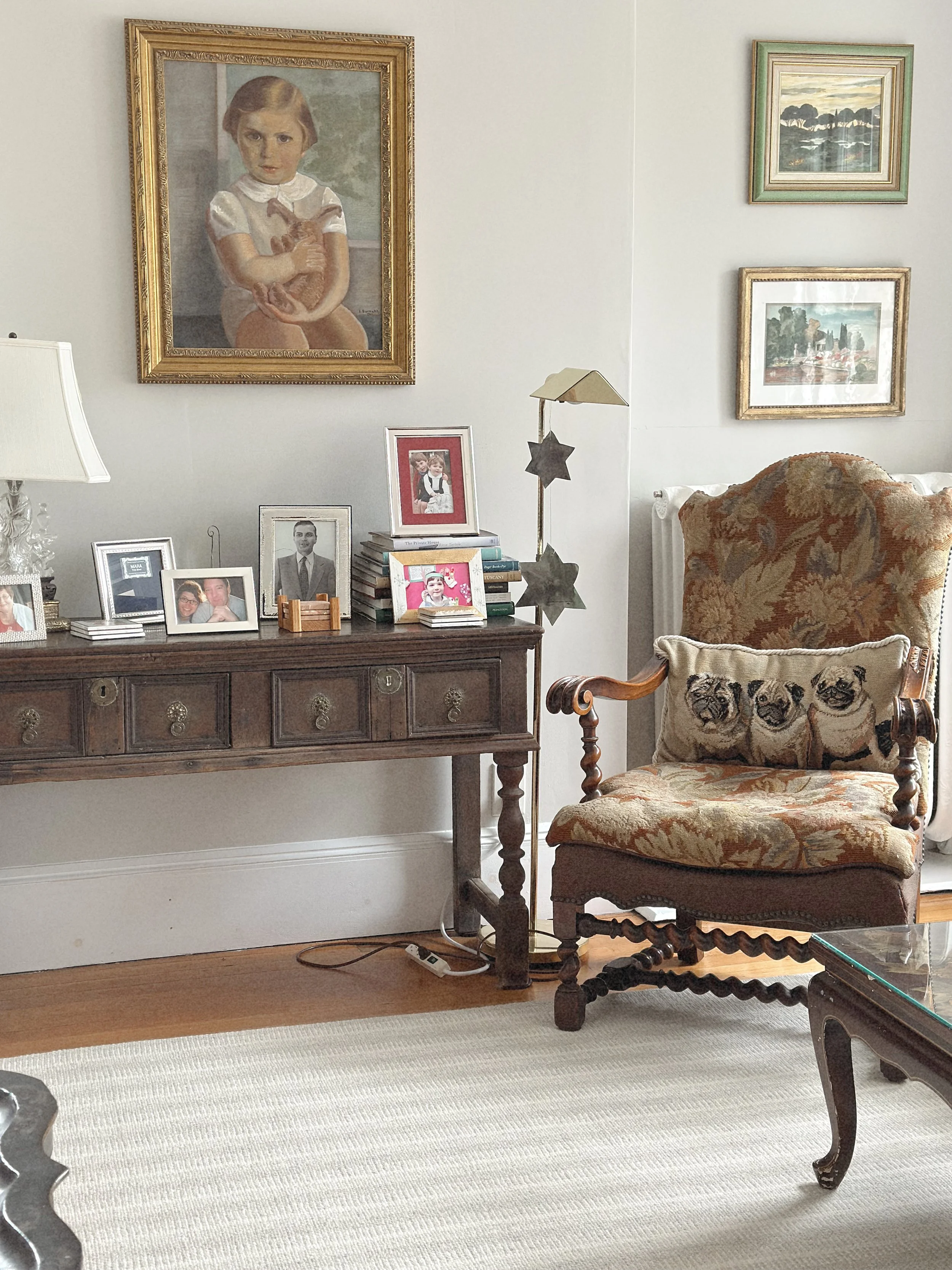 A living room corner with a wooden sideboard topped with family photos, a lamp, and books. An ornate upholstered armchair with pug face pillows sits beside a glass table. The walls display framed artwork and photographs, creating a cozy, traditional 