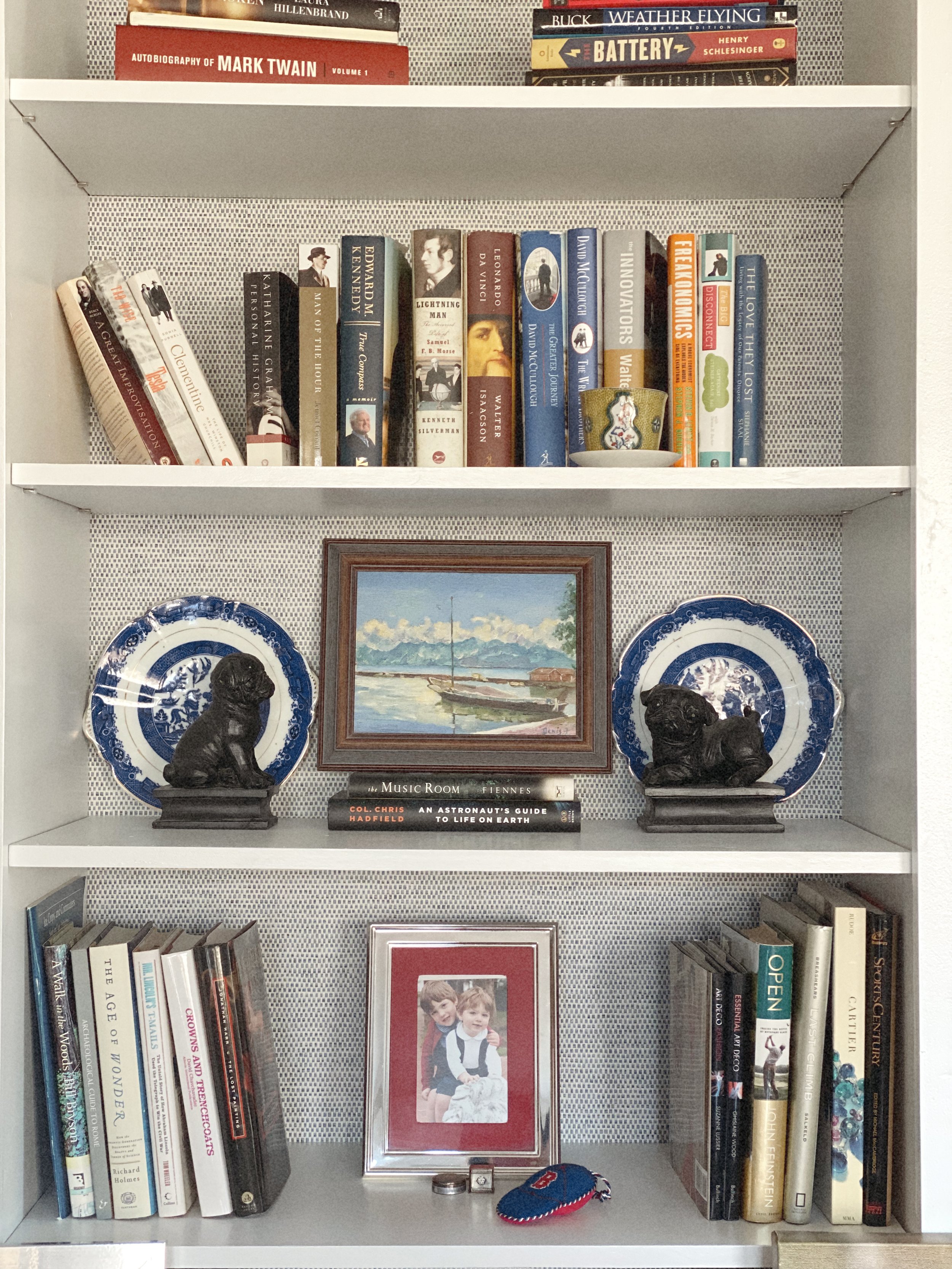 Bookshelf with three shelves containing books, decorative plates, two dog bookends, a framed photo of children, a small decorative tea cup, a key, and a miniature baseball, with some books laid flat and others standing upright.