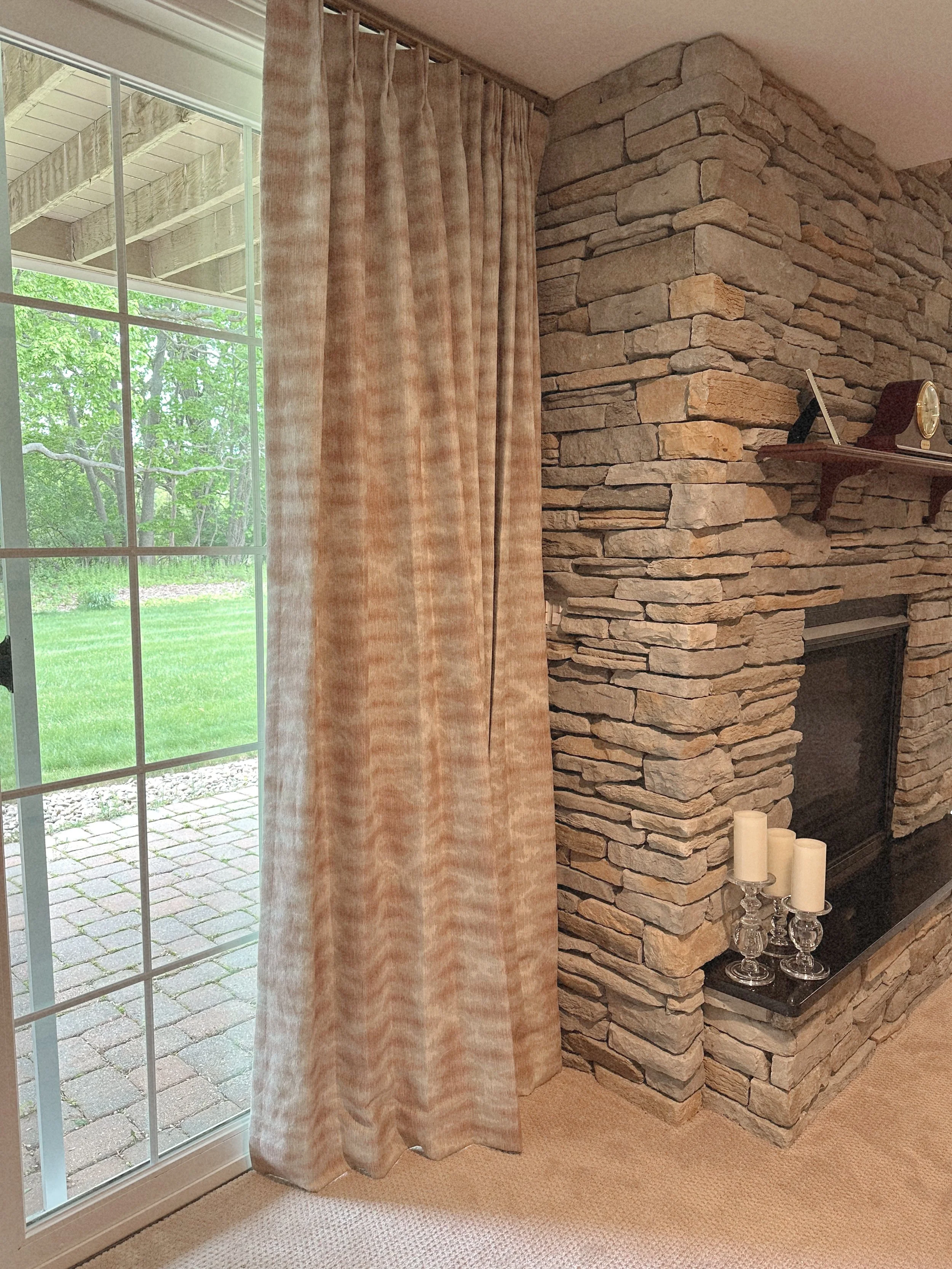 Photo of a living room corner with a stone fireplace, beige curtain, sliding glass door showing an outdoor patio and green yard, and beige carpet.