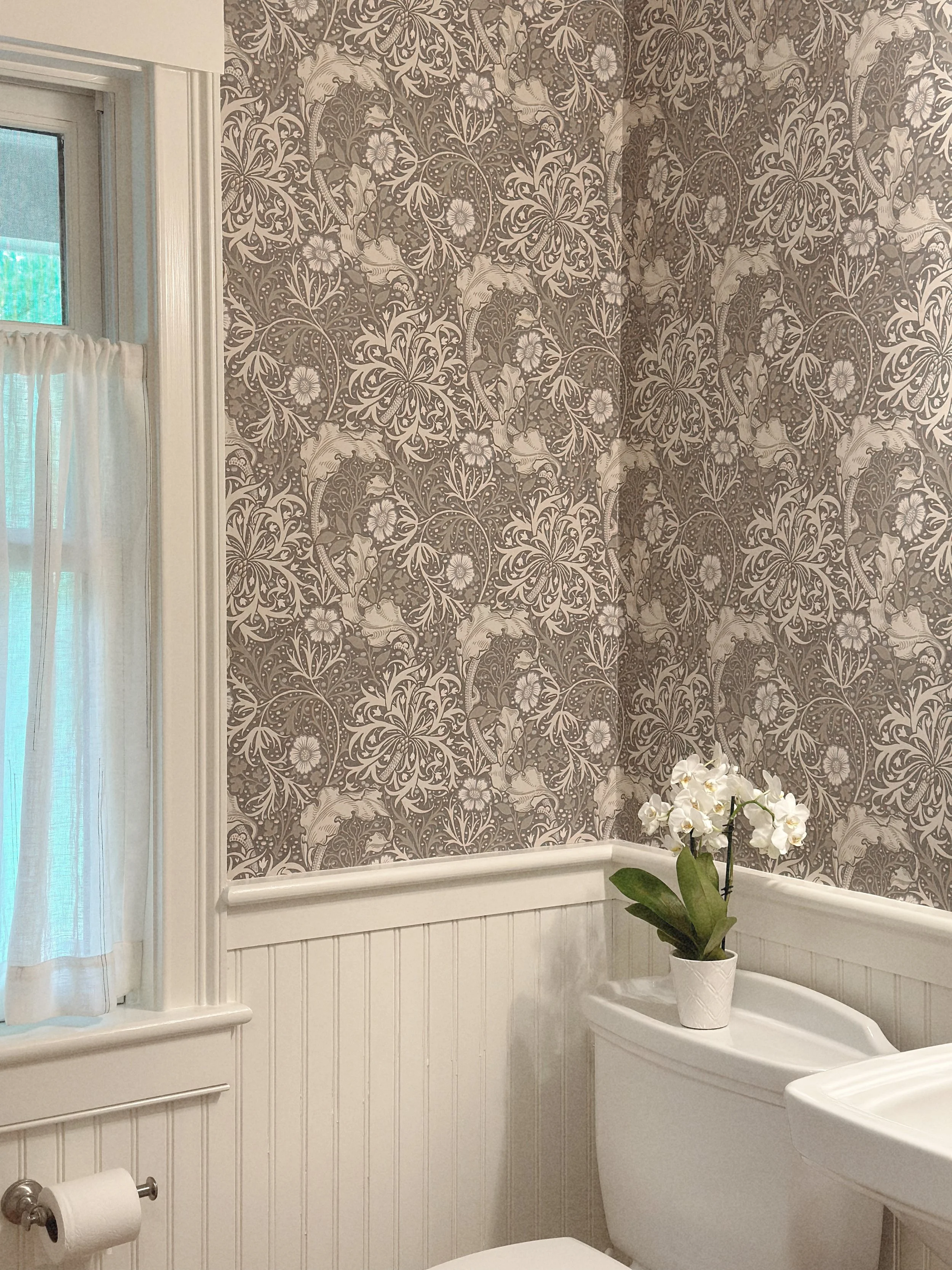 A bathroom with patterned wallpaper featuring floral and animal motifs, a white toilet with a white orchid in a pot on top, a window with a white curtain, and beadboard wainscoting.