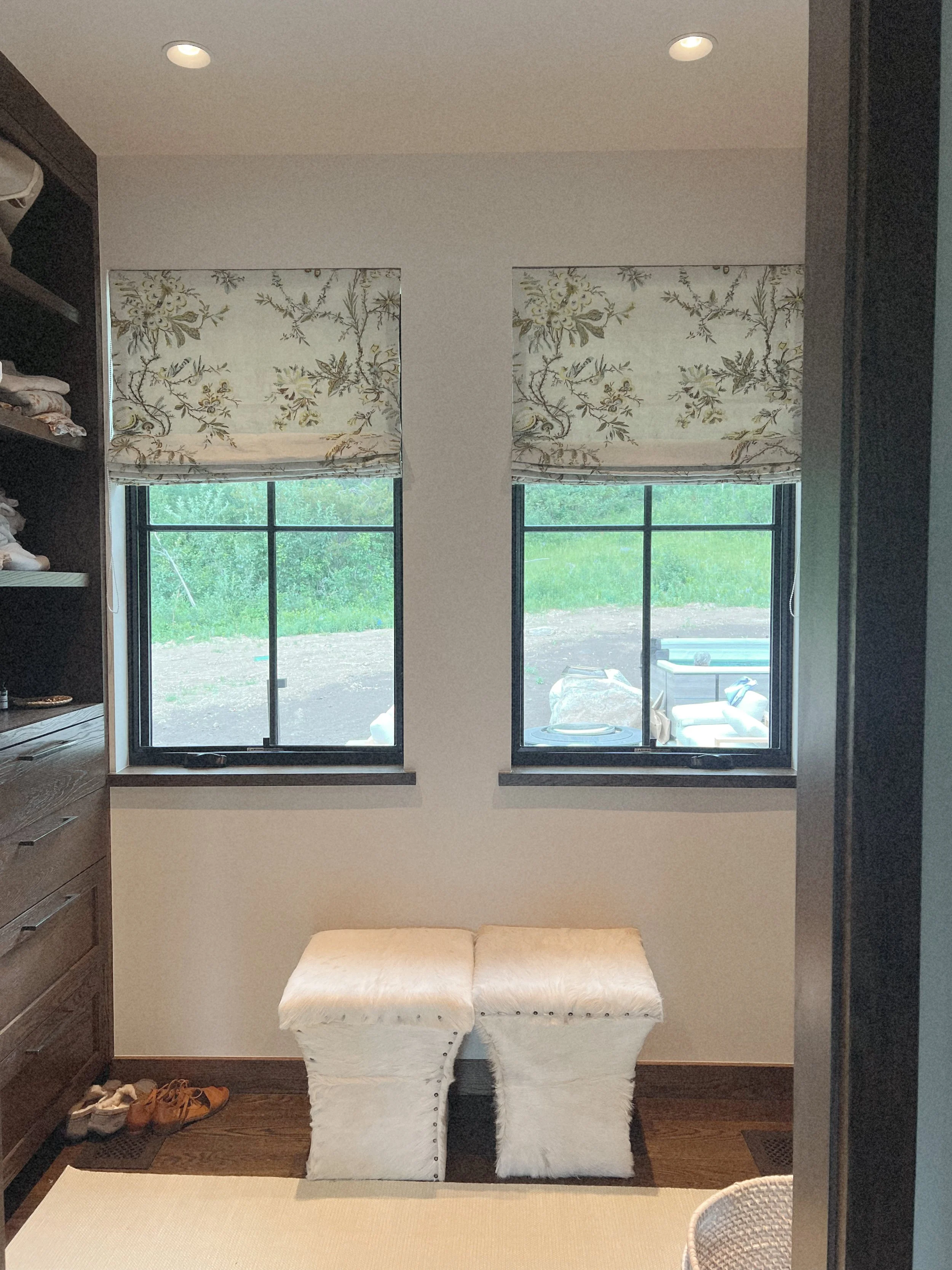 A room with two side-by-side windows with floral-patterned Roman shades, a cream-colored bench with nailhead trim in front, dark wood flooring, a built-in dark wood cabinet with shelves on the left, and a view of a backyard with a hot tub and outdoor