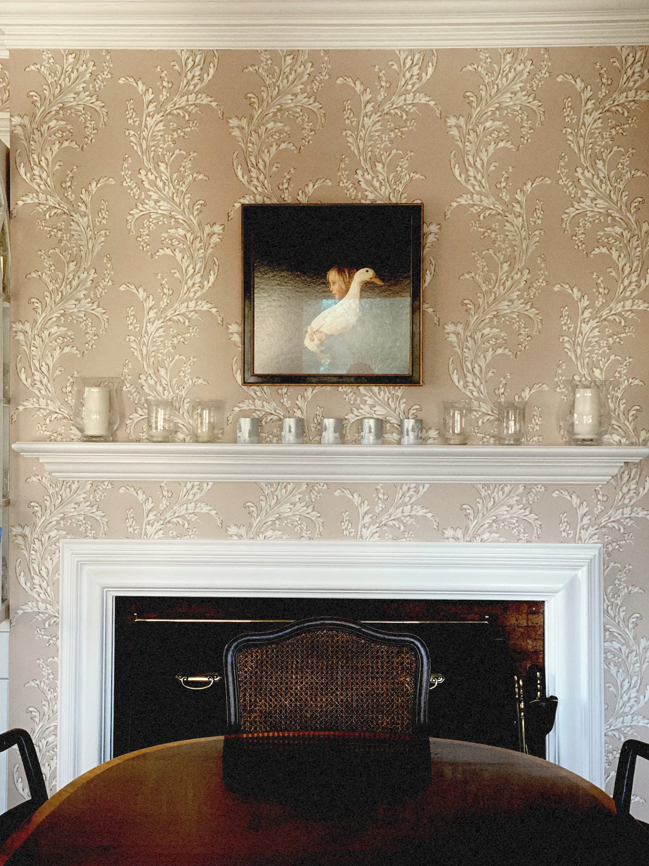 Interior view of a decorated living room with patterned wallpaper, a fireplace with a white mantel, a framed picture of a woman with a boat, a shelf with candles, and a round table with a black bag on it.