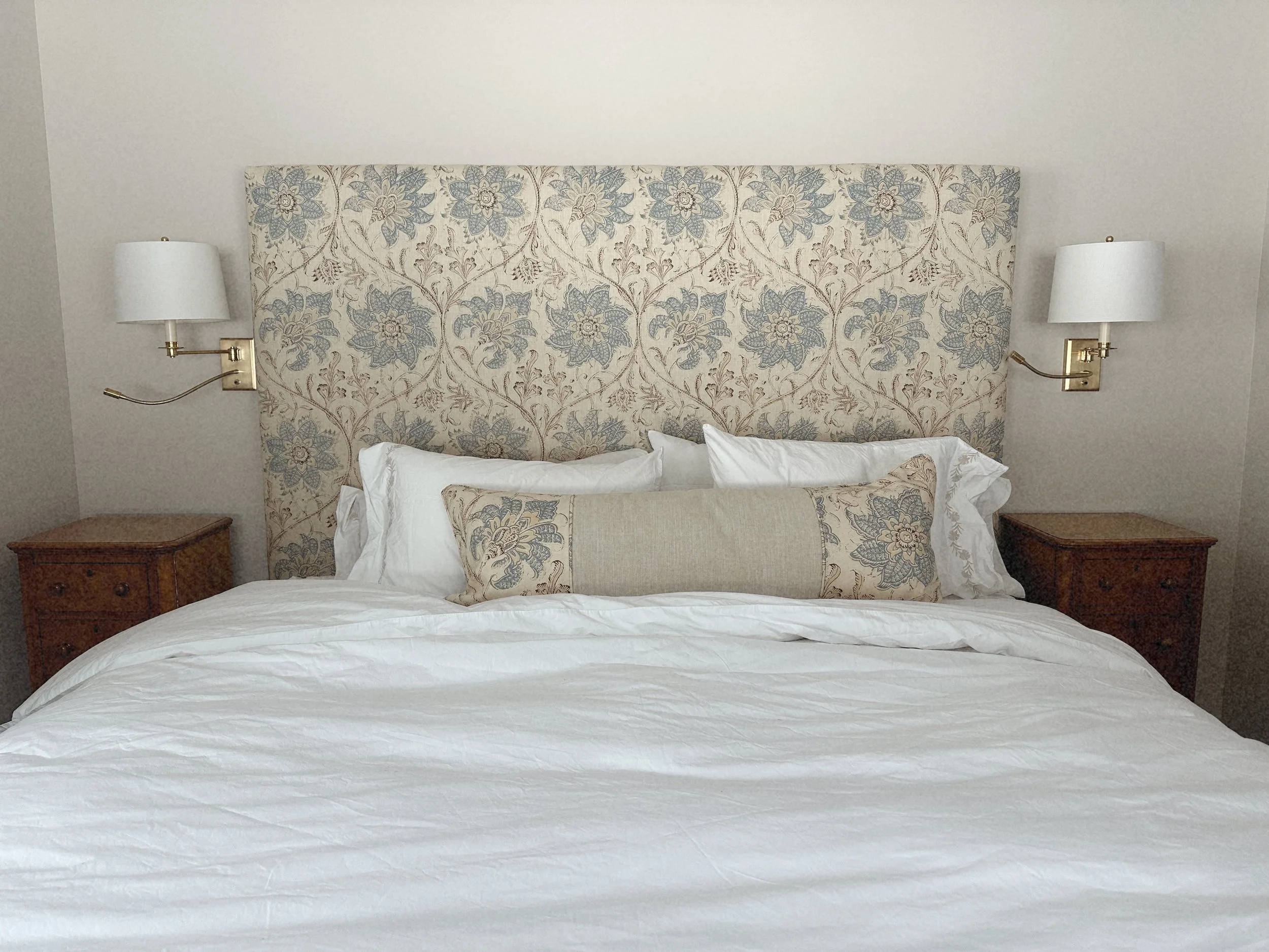A neatly made bed with white linens, a large fabric headboard with floral pattern, and decorative pillows in front. There are two wooden nightstands with lamps on each side of the bed.