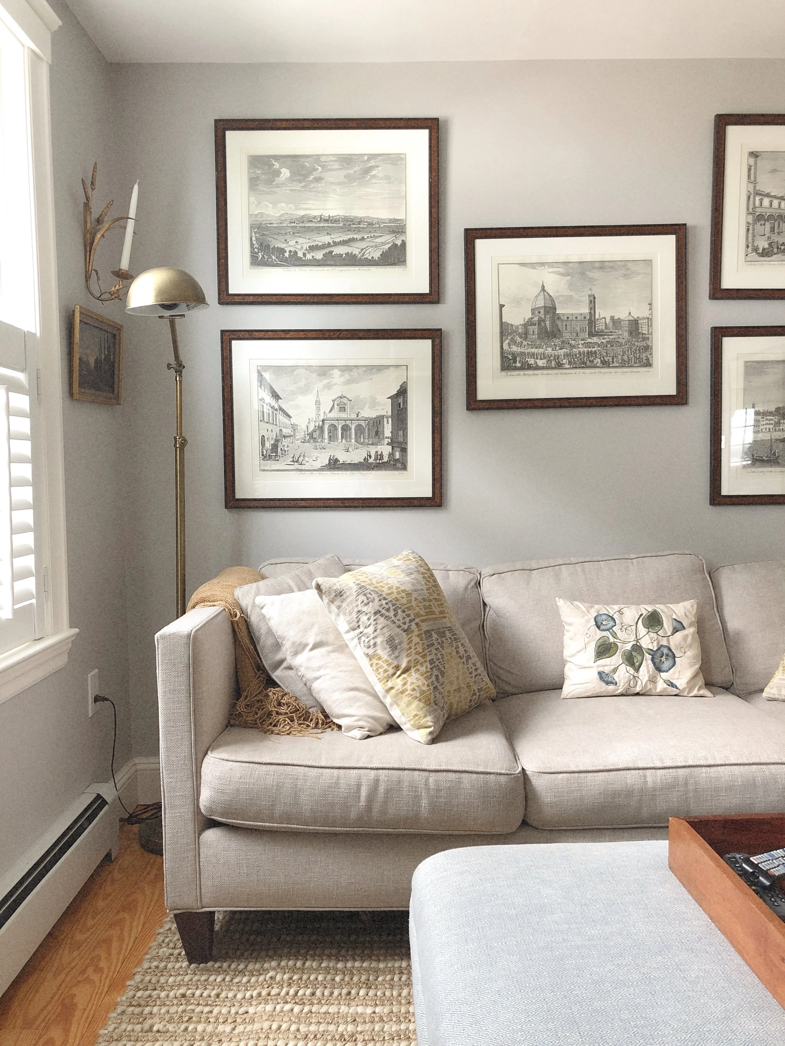 Living room with a beige sofa, decorative pillows, art print wall hangings, a tall floor lamp, and a window with shutters.