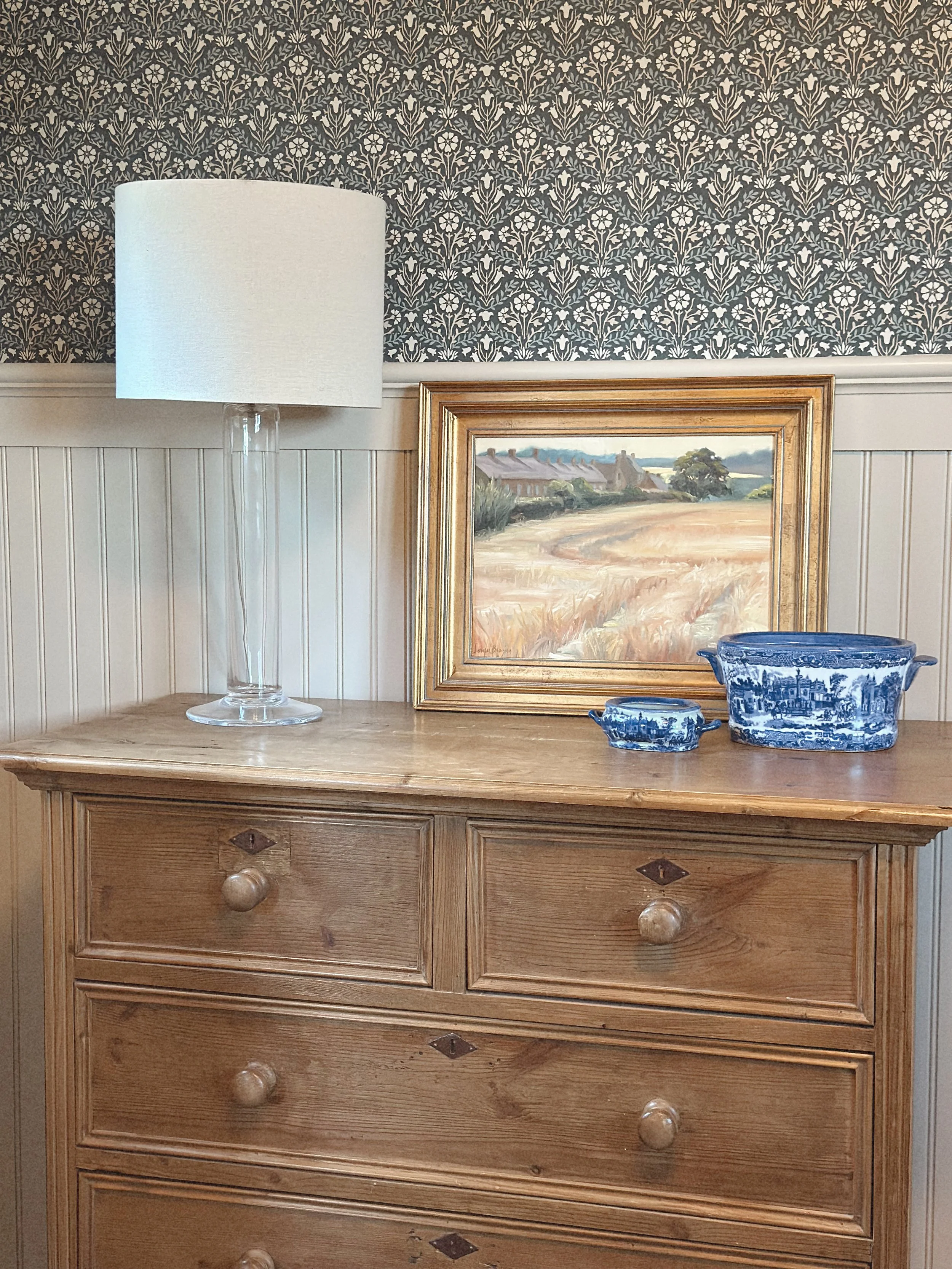 A wooden dresser with a large white lamp, a framed landscape painting, and two blue and white porcelain bowls on top. The background has patterned wallpaper and wood paneling.