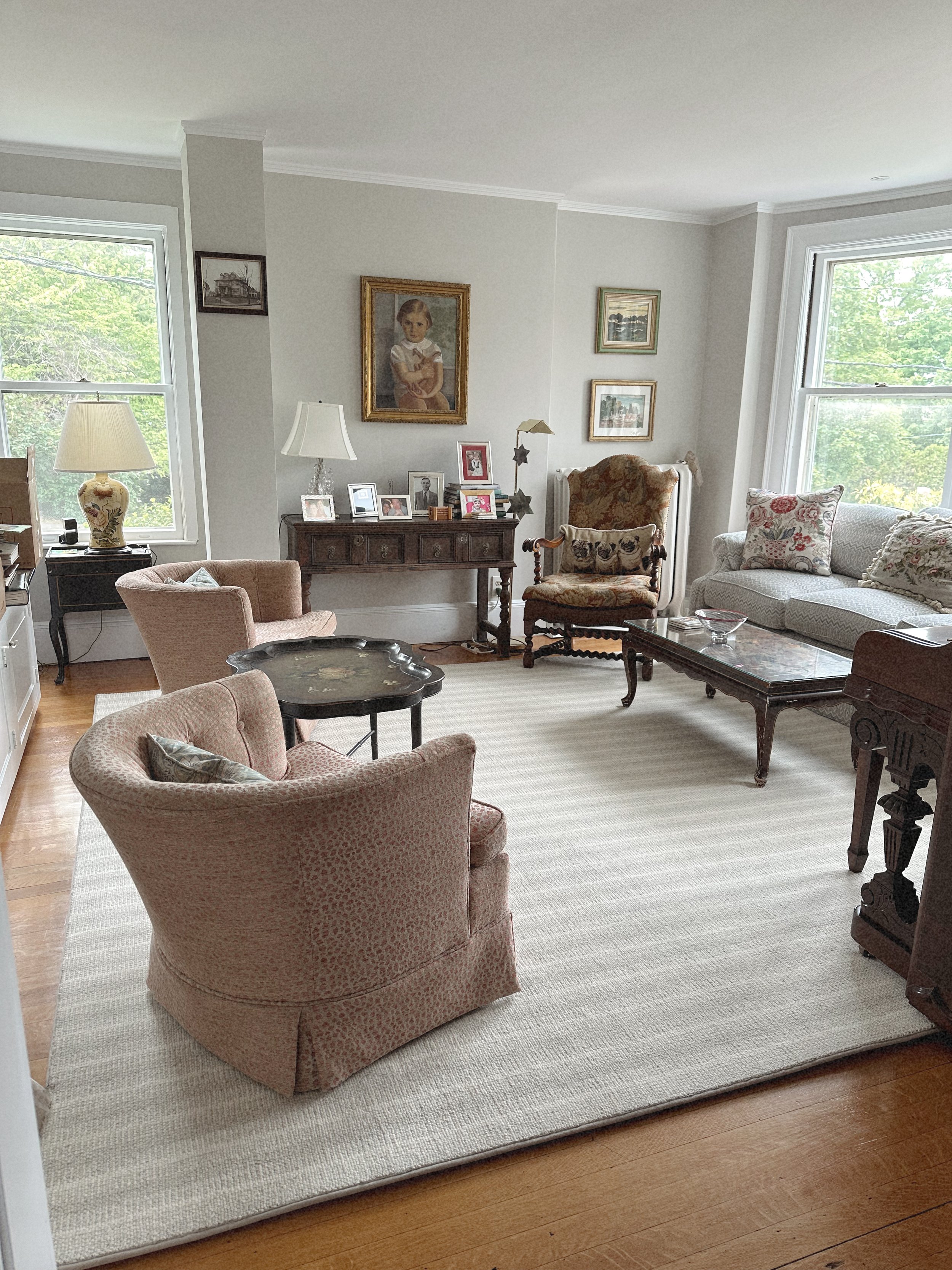 A living room with antique furniture, framed pictures, a beige armchair, a floral sofa, and large windows with greenery outside.